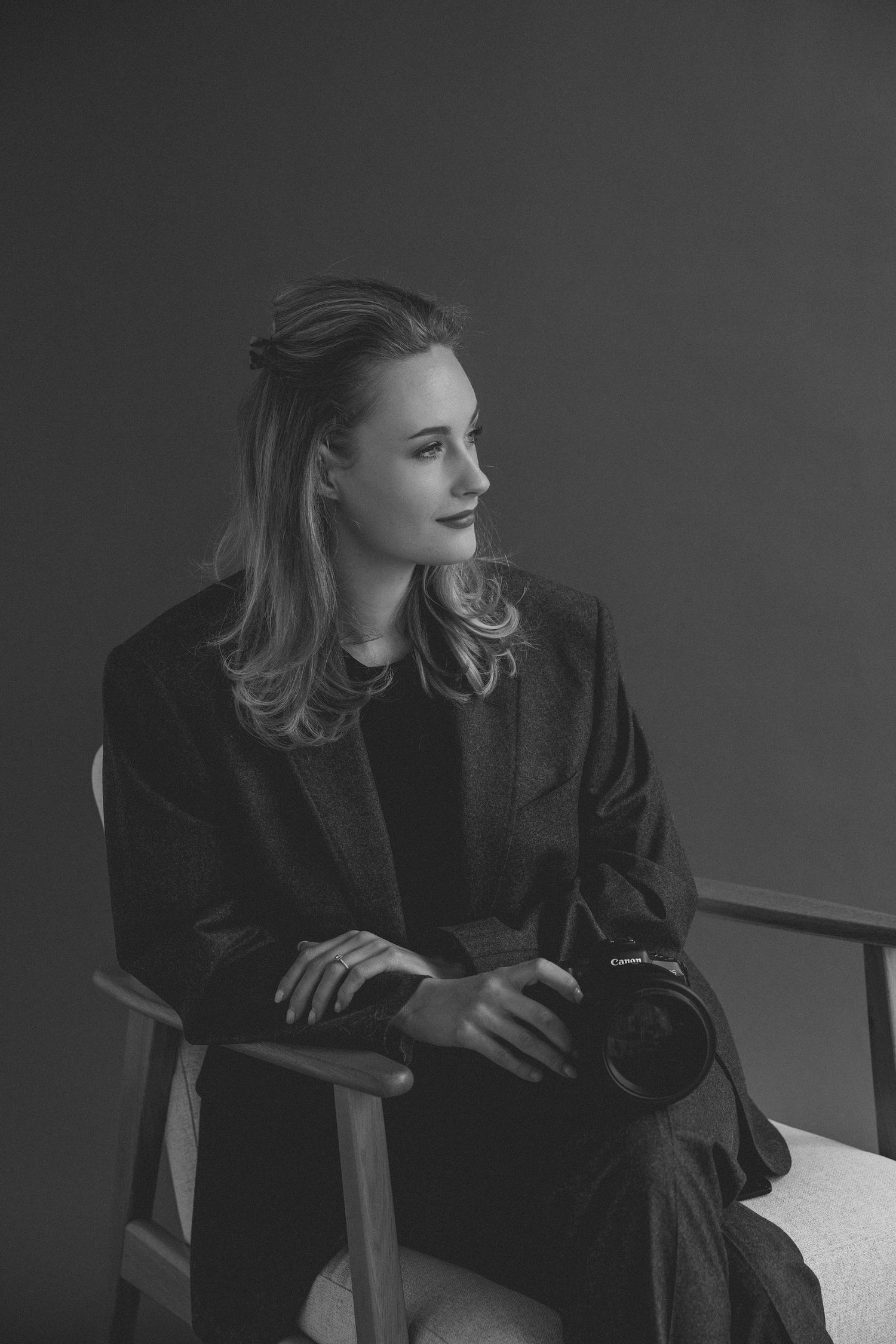 Black and white portrait of a woman in a blazer sitting in a chair holding a Canon camera, looking to the side.