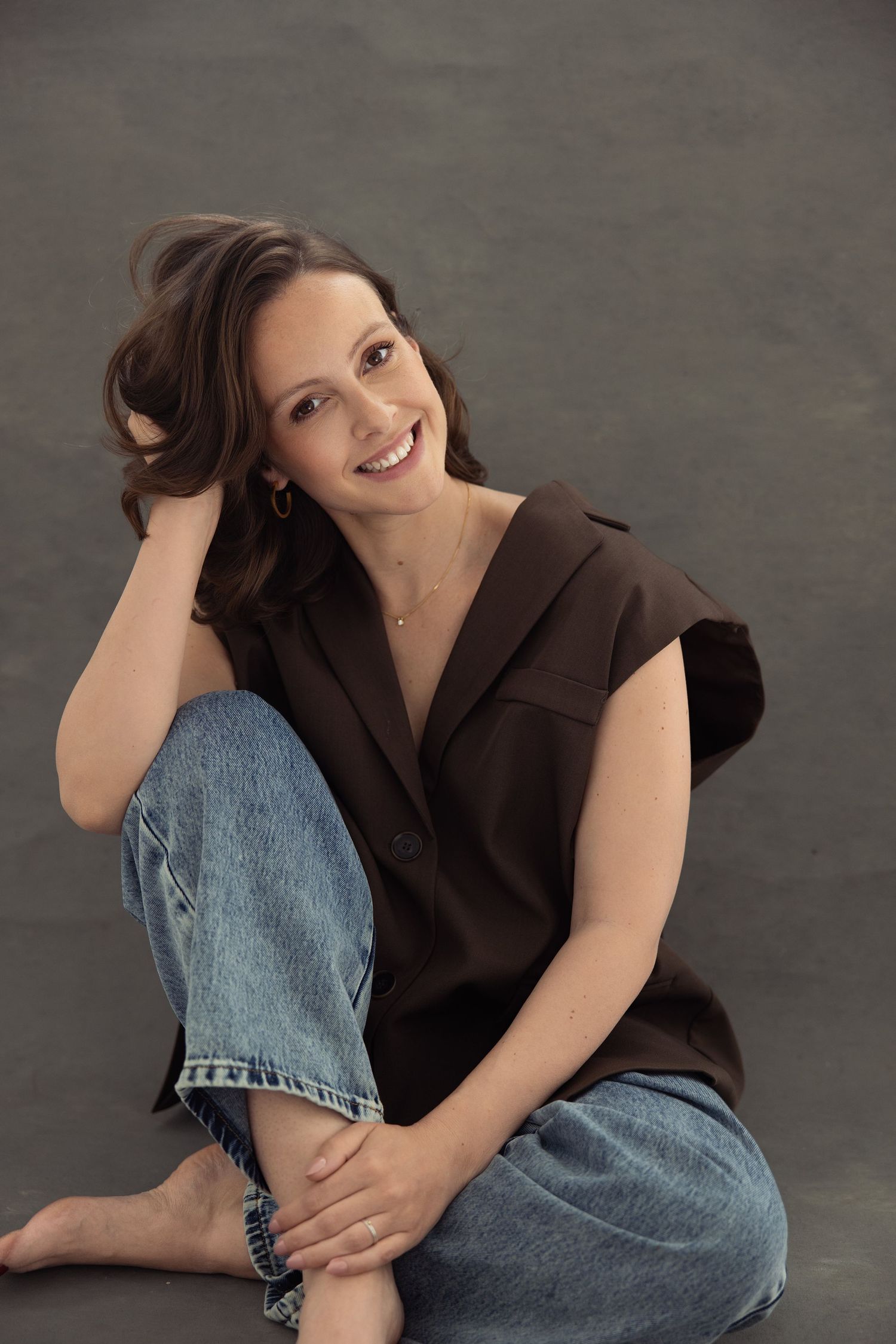 Smiling woman in brown top and jeans sitting relaxed on floor against gray background.
