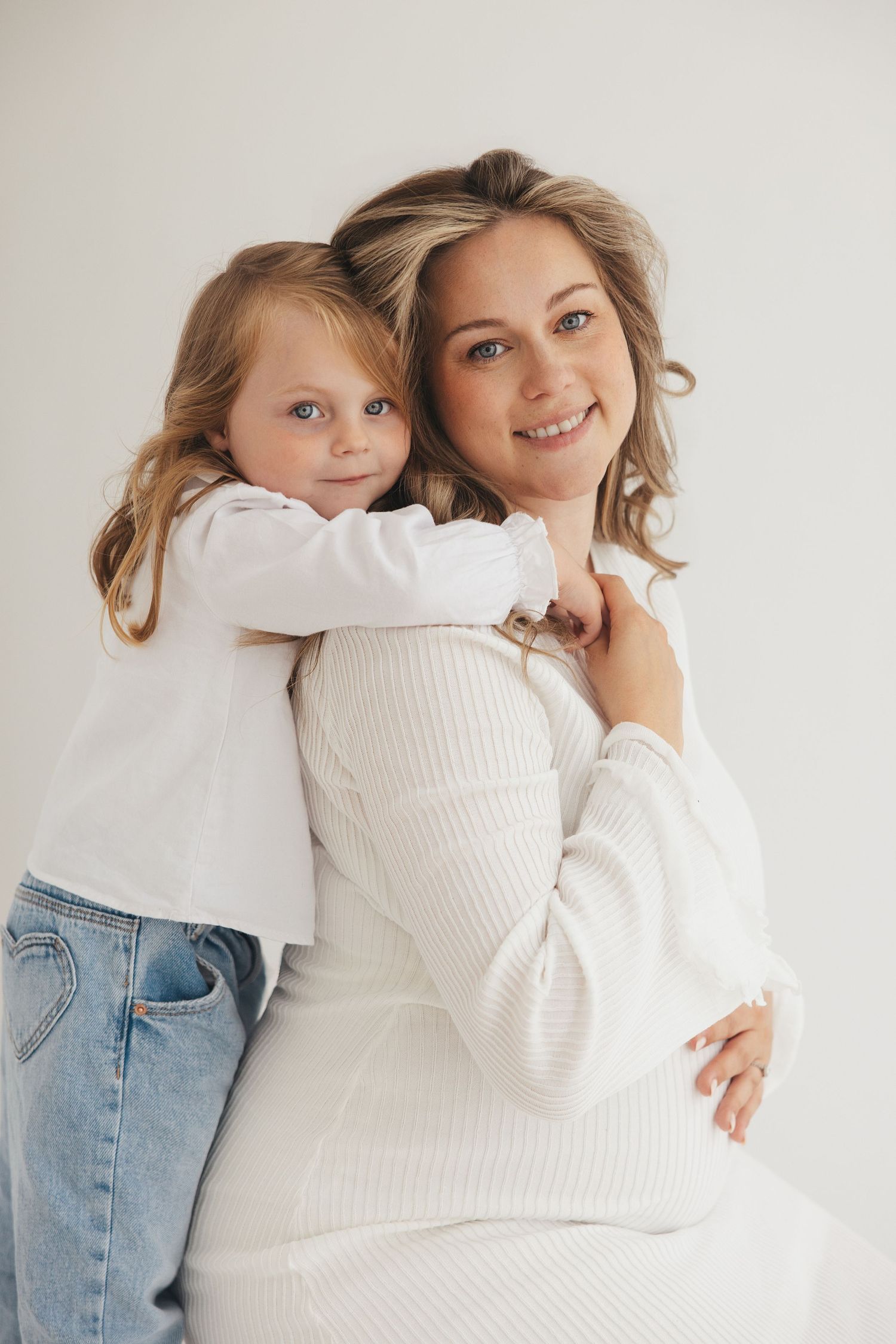 A young girl hugs a smiling blonde woman in white, both with light hair, posed against a white background.