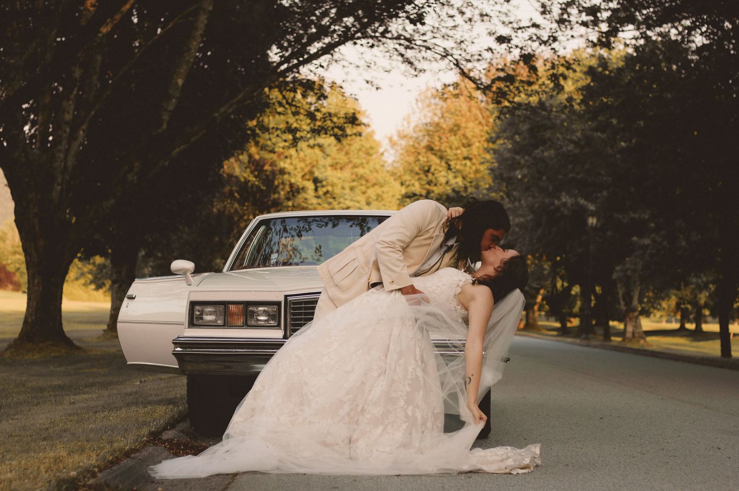 Wedding couple posing by vintage car during golden hour in a tree-lined park.