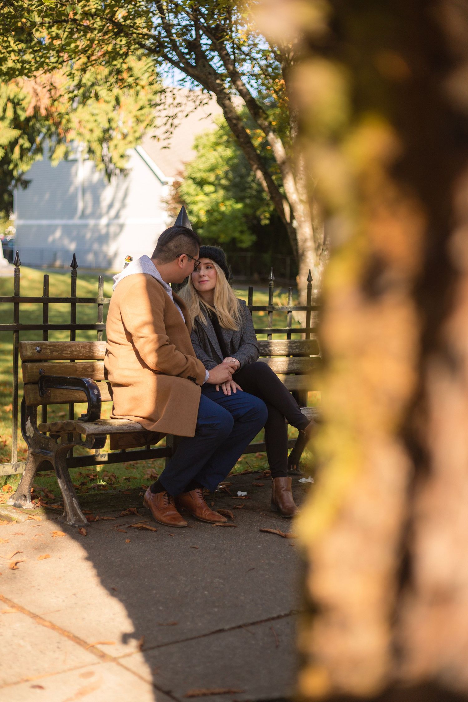 A couple sits close together on a wooden park bench surrounded by autumn trees.