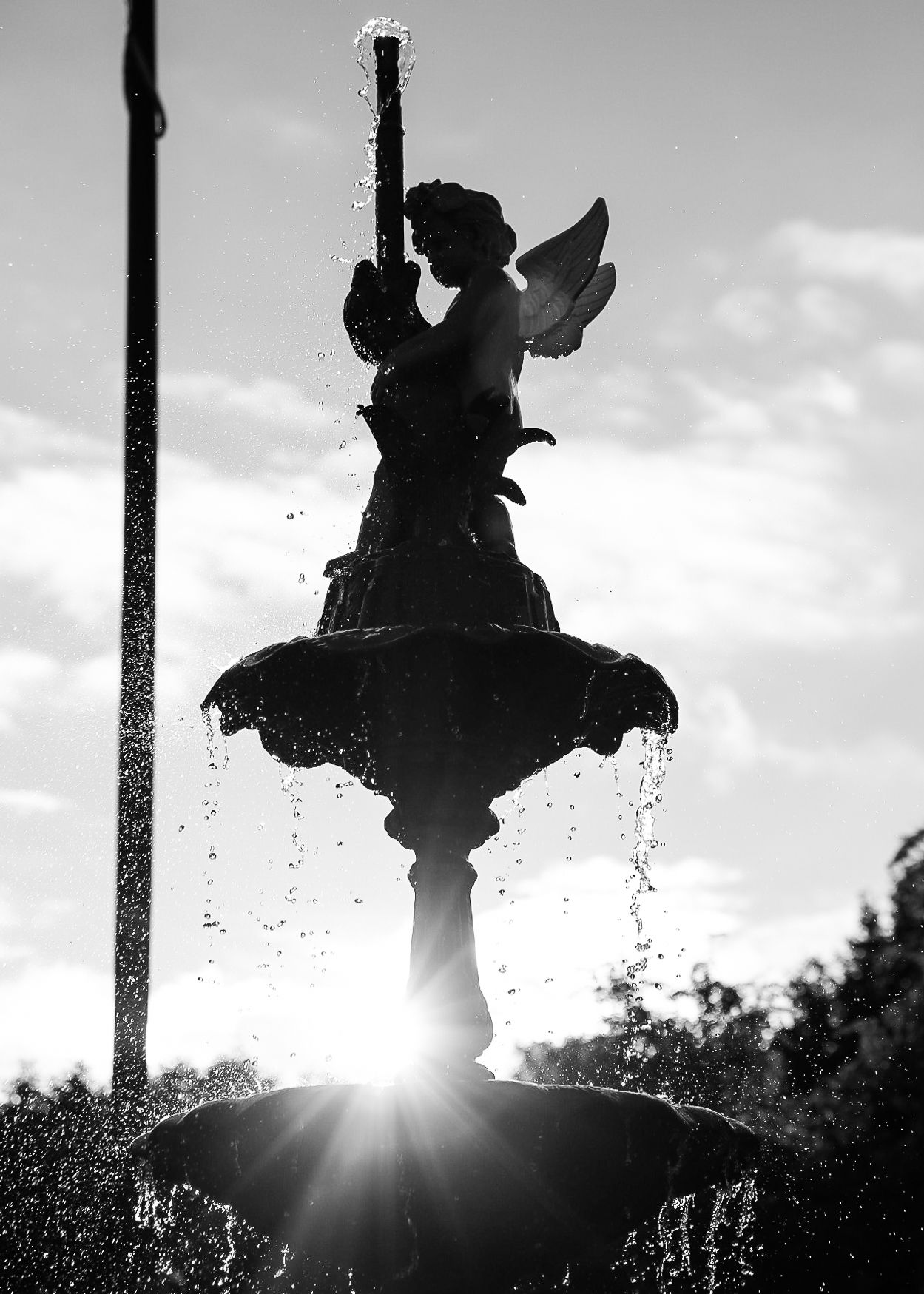 Silhouette of an angel fountain statue against bright sunlight with water spraying.