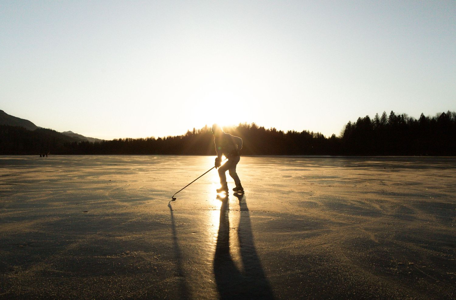 Silhouetted skaters cast long shadows while ice skating on a frozen lake at sunset with mountains in the background.