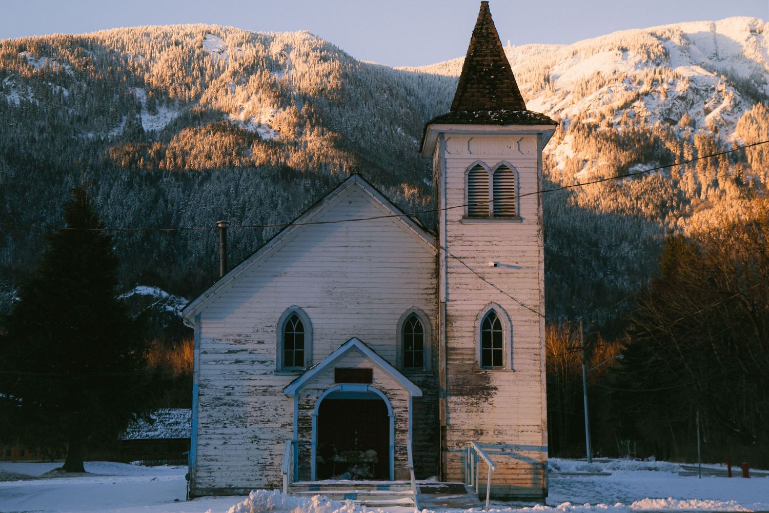 Historic white church with steeple nestled against snow-covered mountains at sunset.