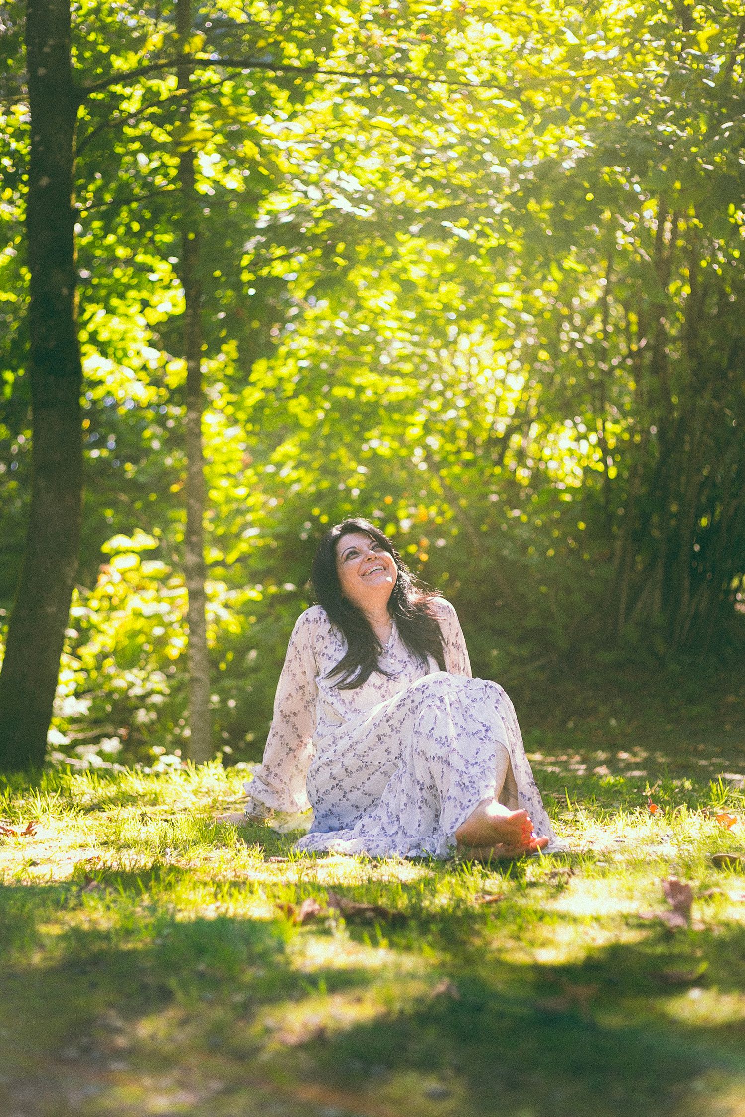 A person in a white lace dress sits on grass in dappled sunlight surrounded by trees.