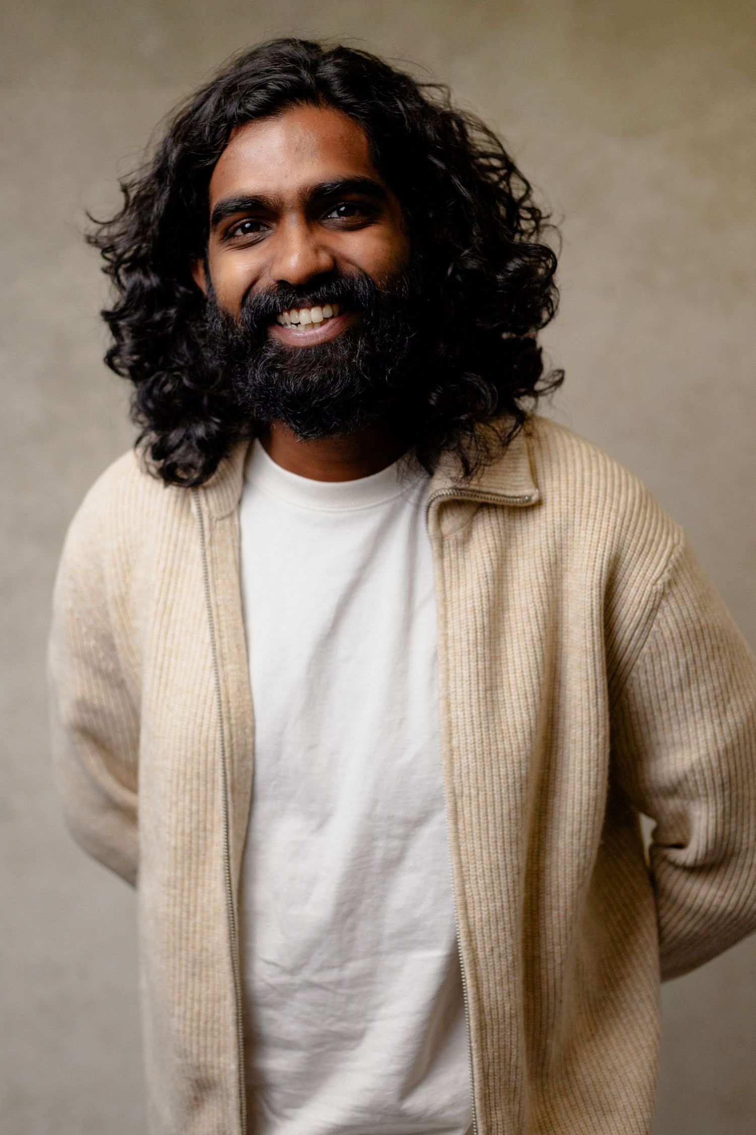 Portrait of a person with long curly dark hair wearing a beige cardigan and white t-shirt against a neutral background.