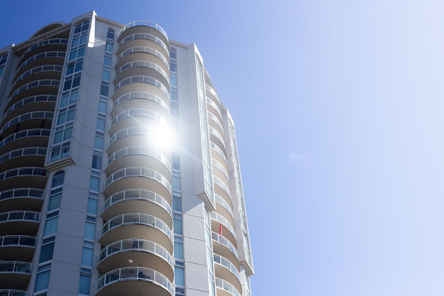 Modern white high-rise condominium building reflecting bright sunlight against blue sky.