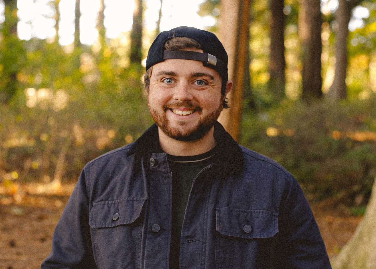 Casual portrait of someone wearing a backwards cap and denim jacket in a wooded setting.