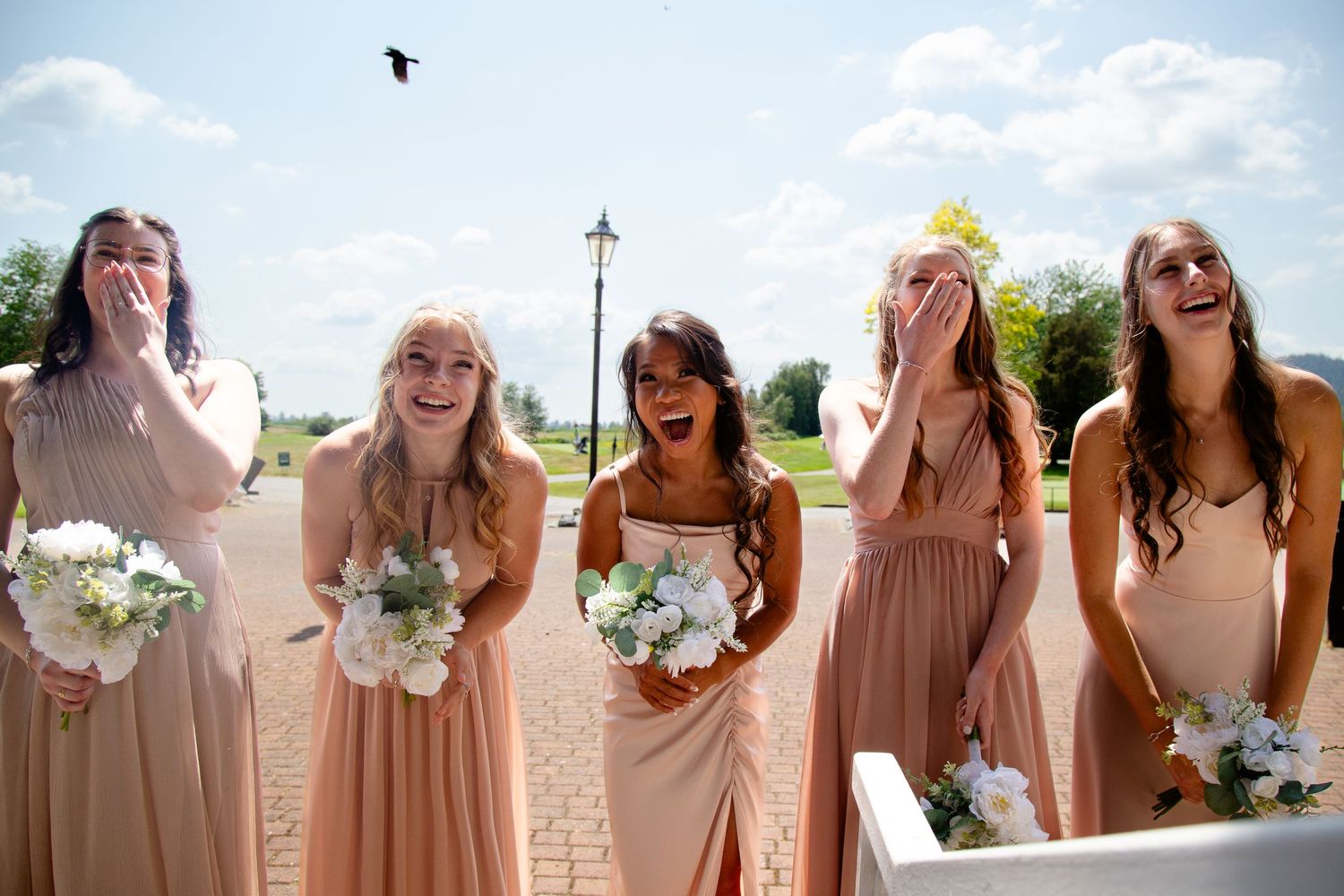 Bridesmaids in peach dresses holding white bouquets share a candid laughing moment.