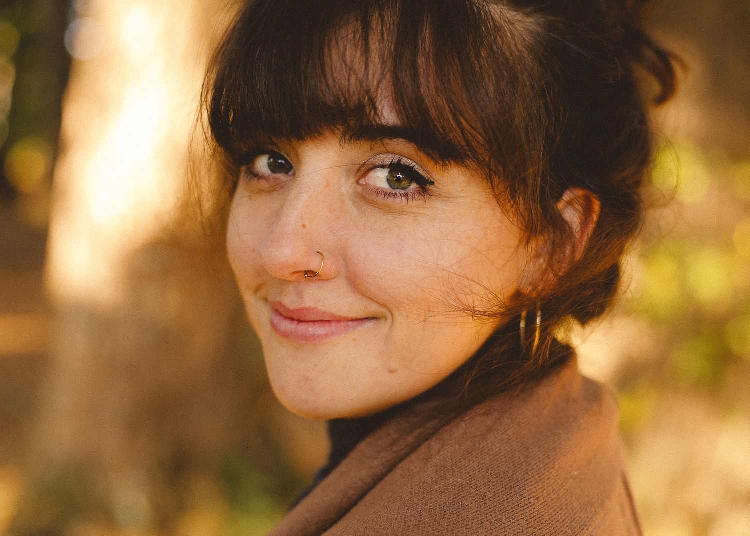 A portrait in warm lighting showing someone wearing a brown coat and bangs.