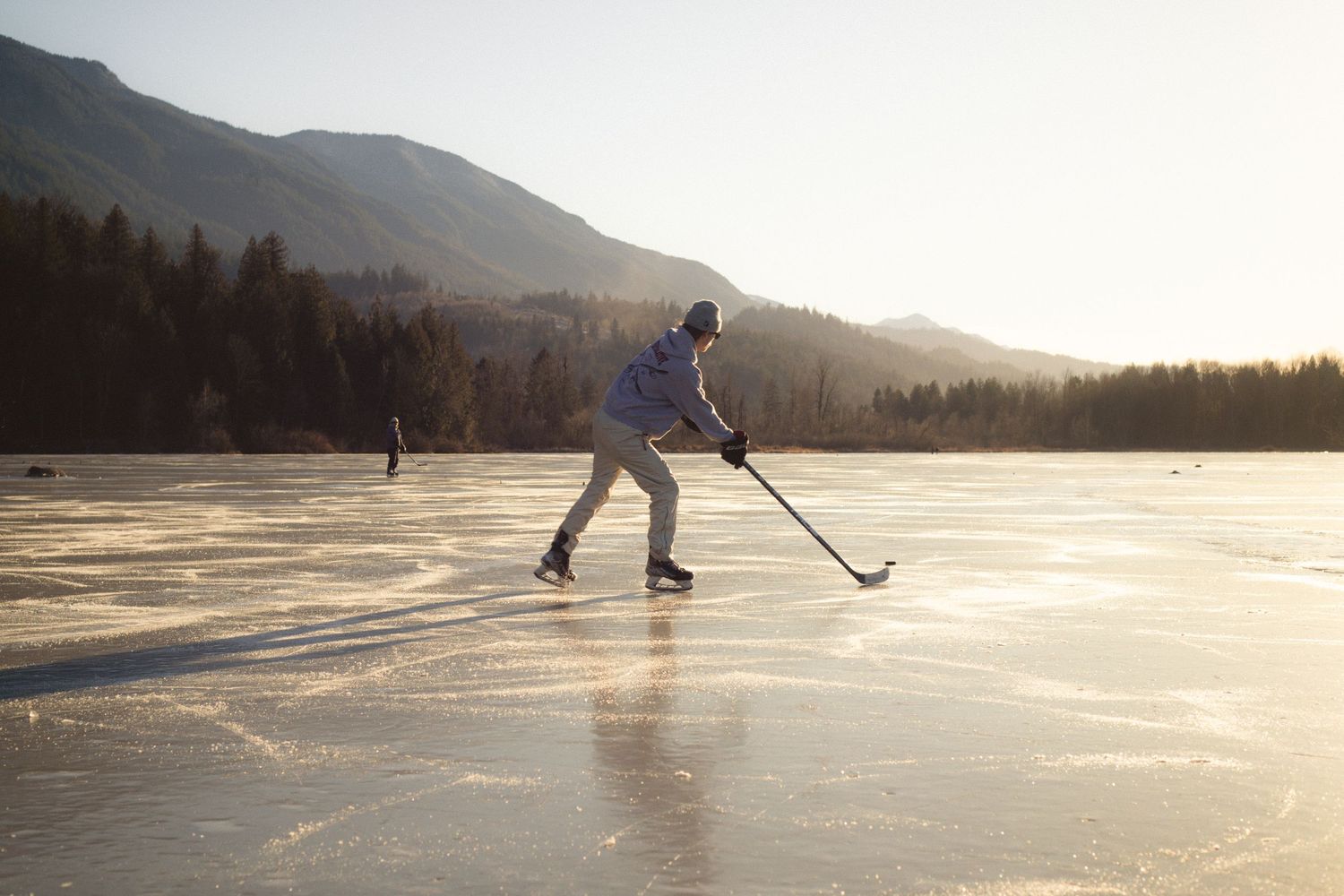 A hockey player practices on a frozen mountain lake at sunset with mountains and forest in the background.
