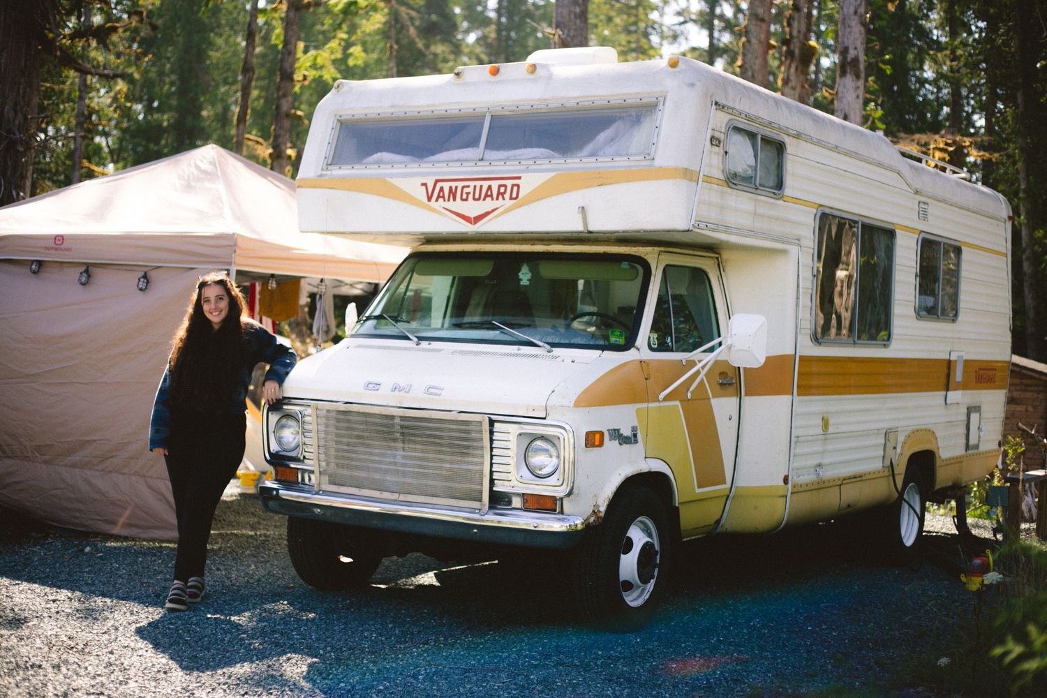 Vintage RV camper van parked in a wooded campsite with awning extended.
