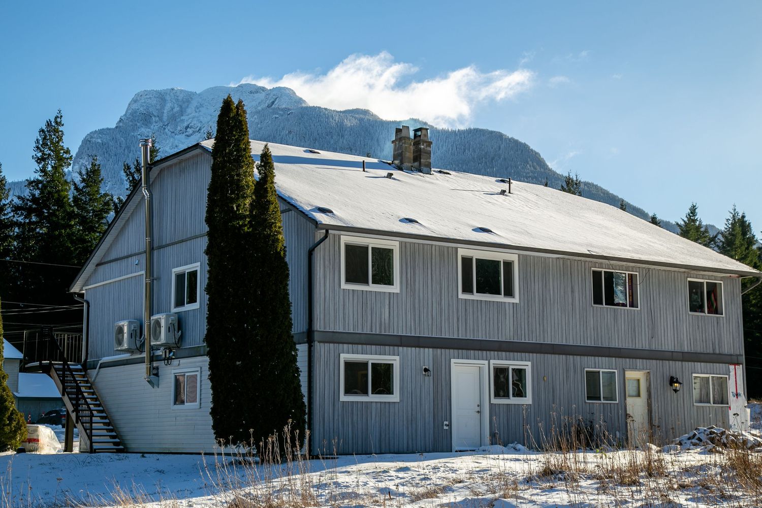 Grey two-story building with snow on its roof against a mountainous winter backdrop.