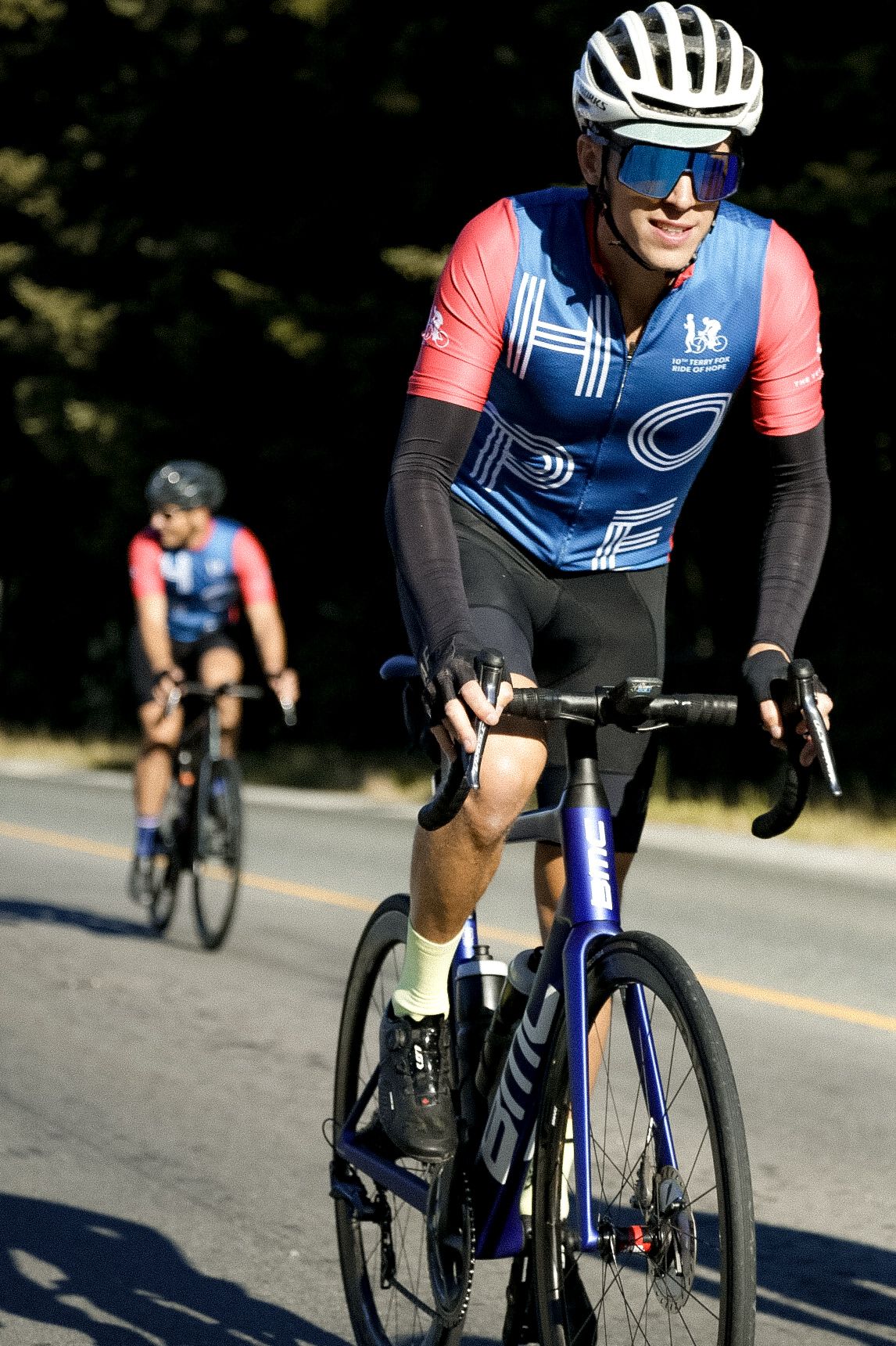 Cyclist wearing blue and pink jersey rides road bike on sunny asphalt road with another rider in background.