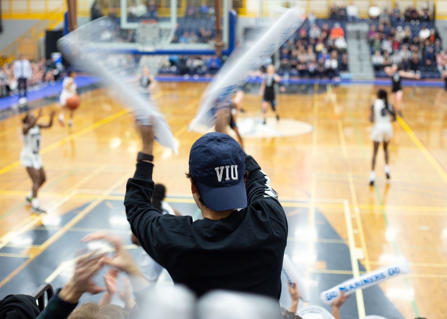 Spectator wearing a VIU cap watching an indoor basketball game from the bleachers.