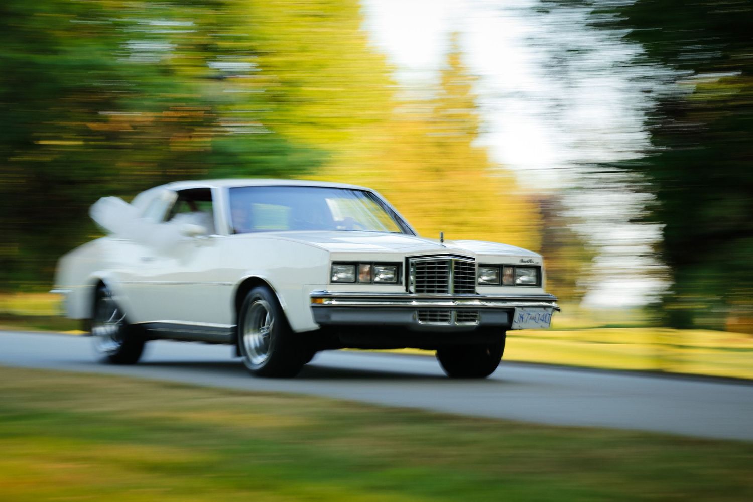 A classic white Mercury Cougar car in motion on a tree-lined road with motion blur effect.