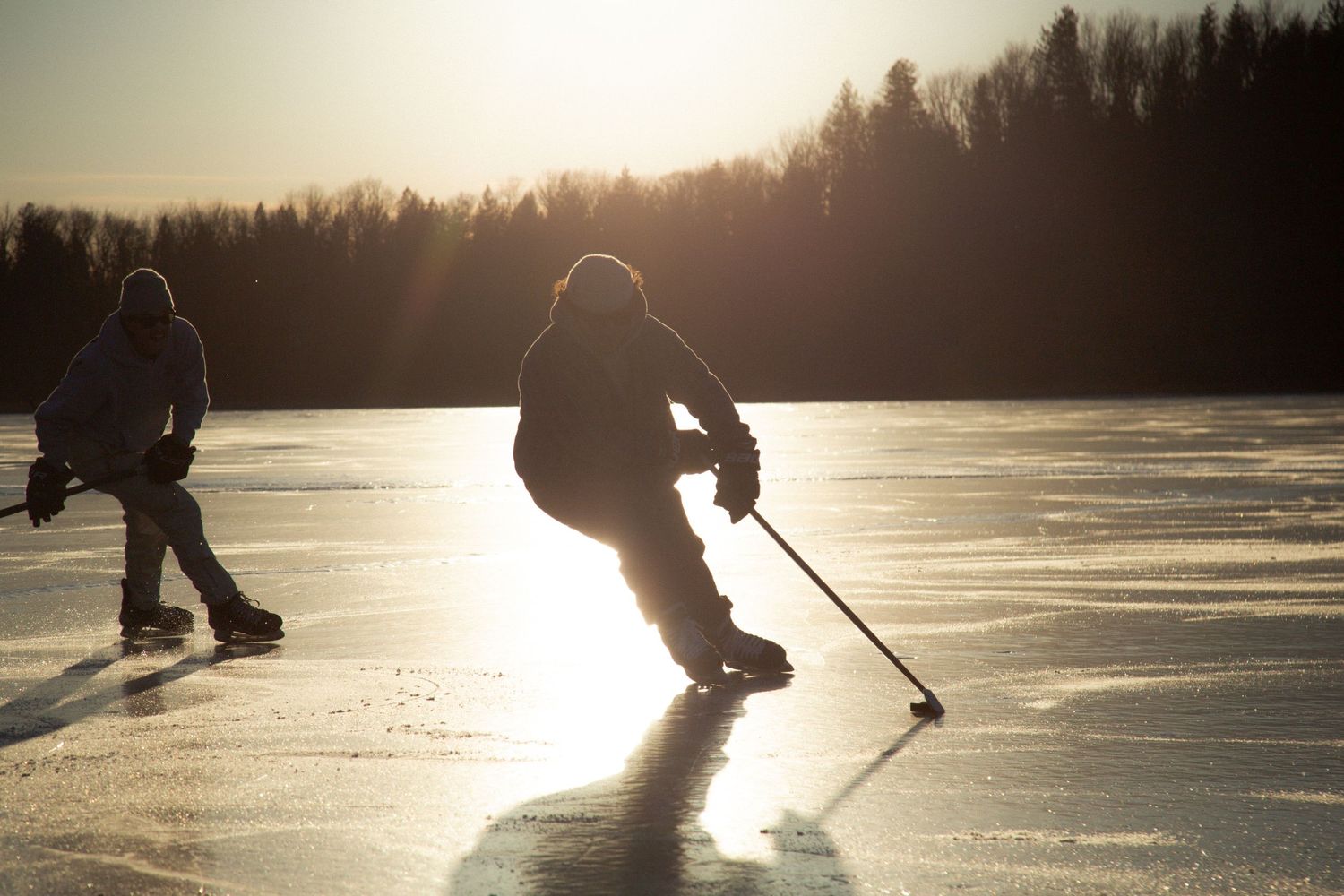Silhouettes of hockey players on a frozen lake during sunset with winter forest backdrop casting long shadows on ice.