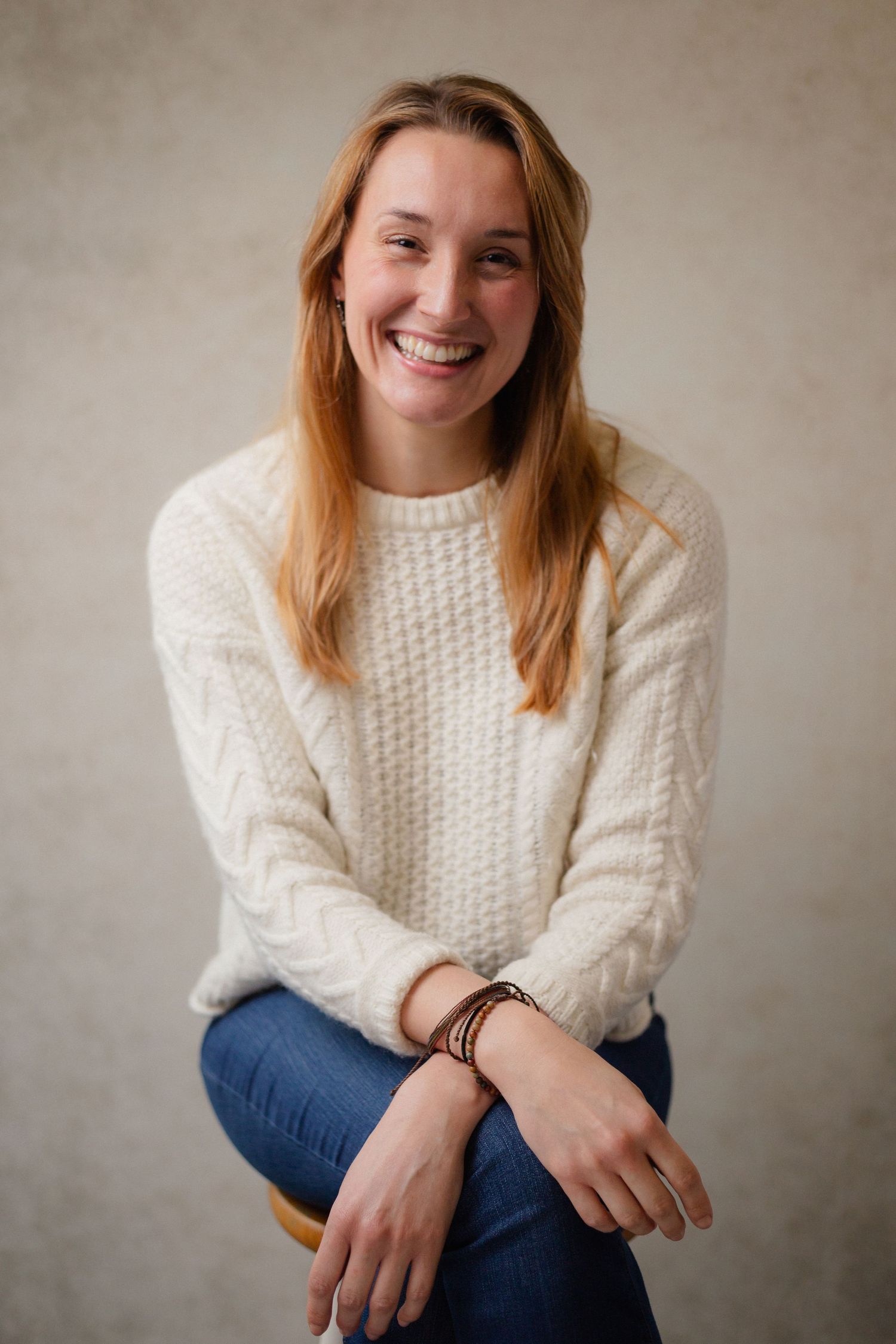 A portrait of someone in a white cable knit sweater and jeans seated casually.