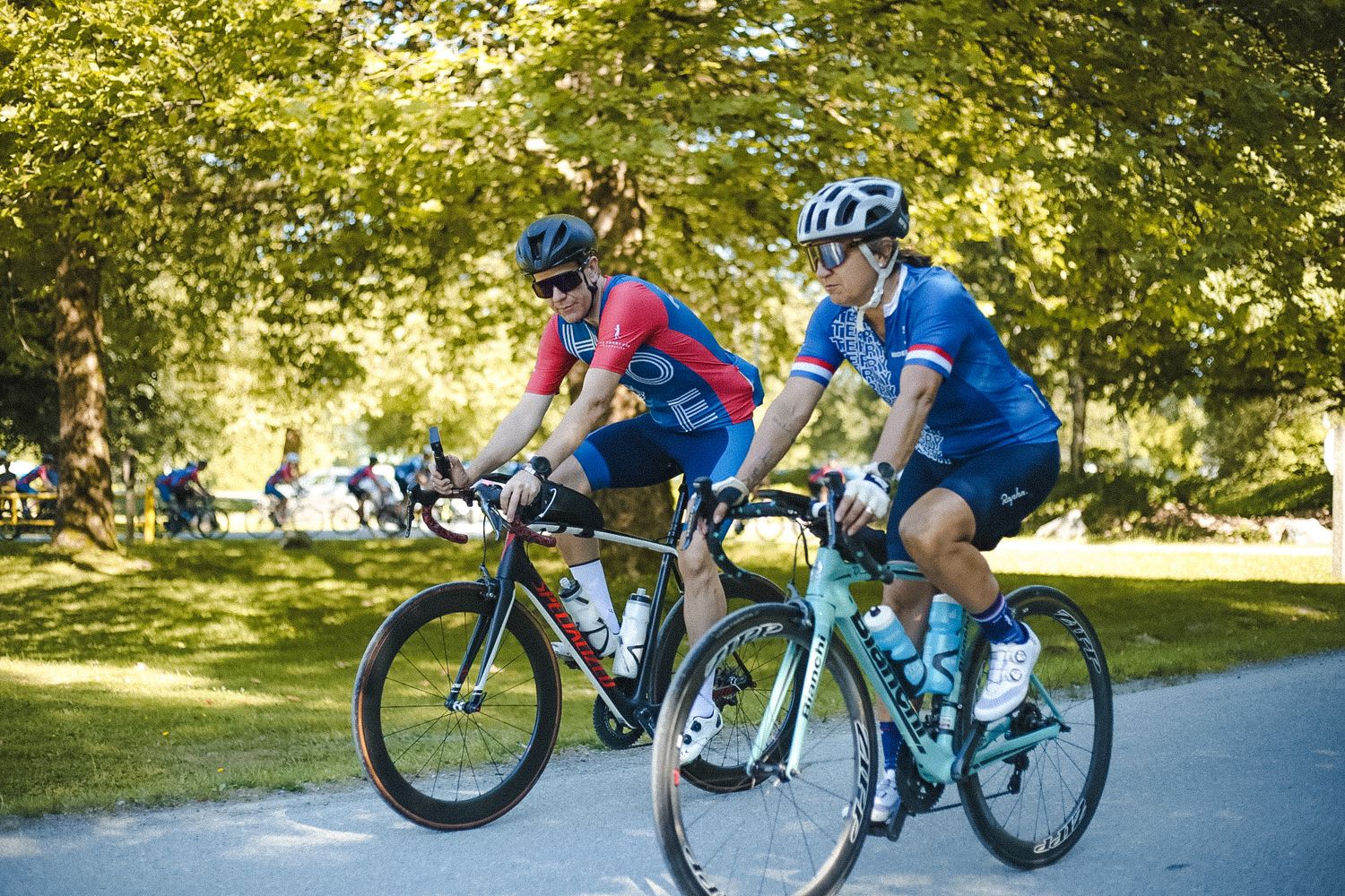 Two cyclists in bright jerseys riding side by side on a tree-shaded path.