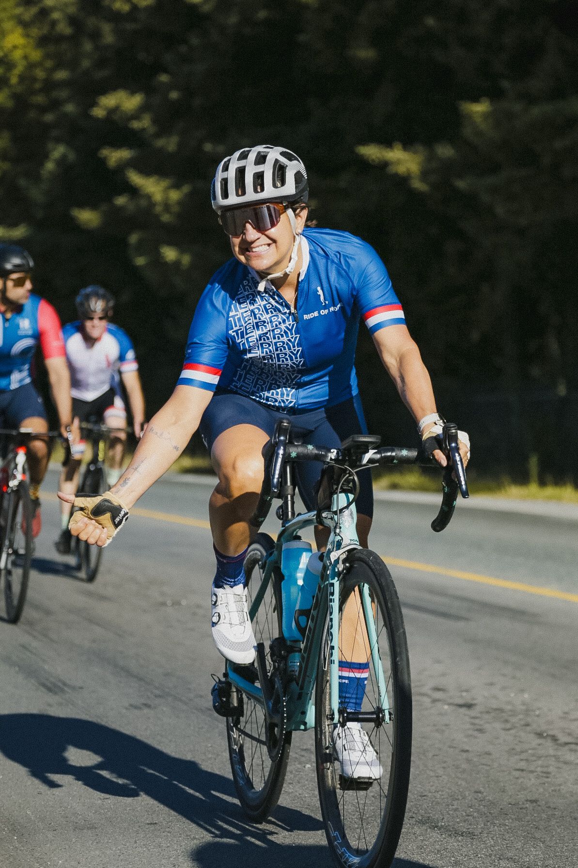 Cyclist wearing blue jersey and helmet rides a road bike on a sunny day with other riders in the background.
