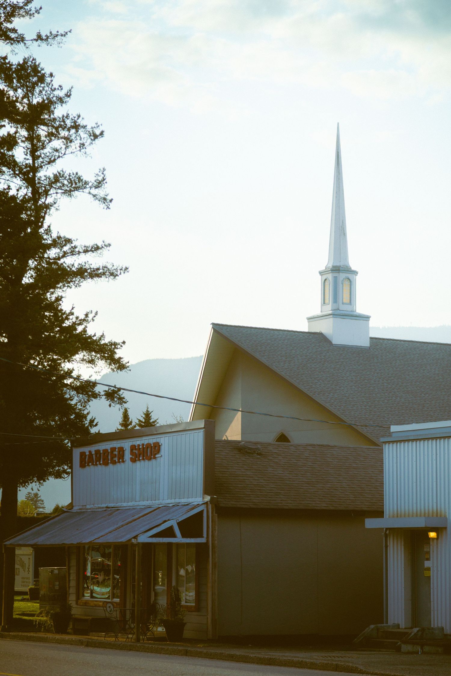 A small barber shop storefront with a white church steeple rising in the background during golden hour.