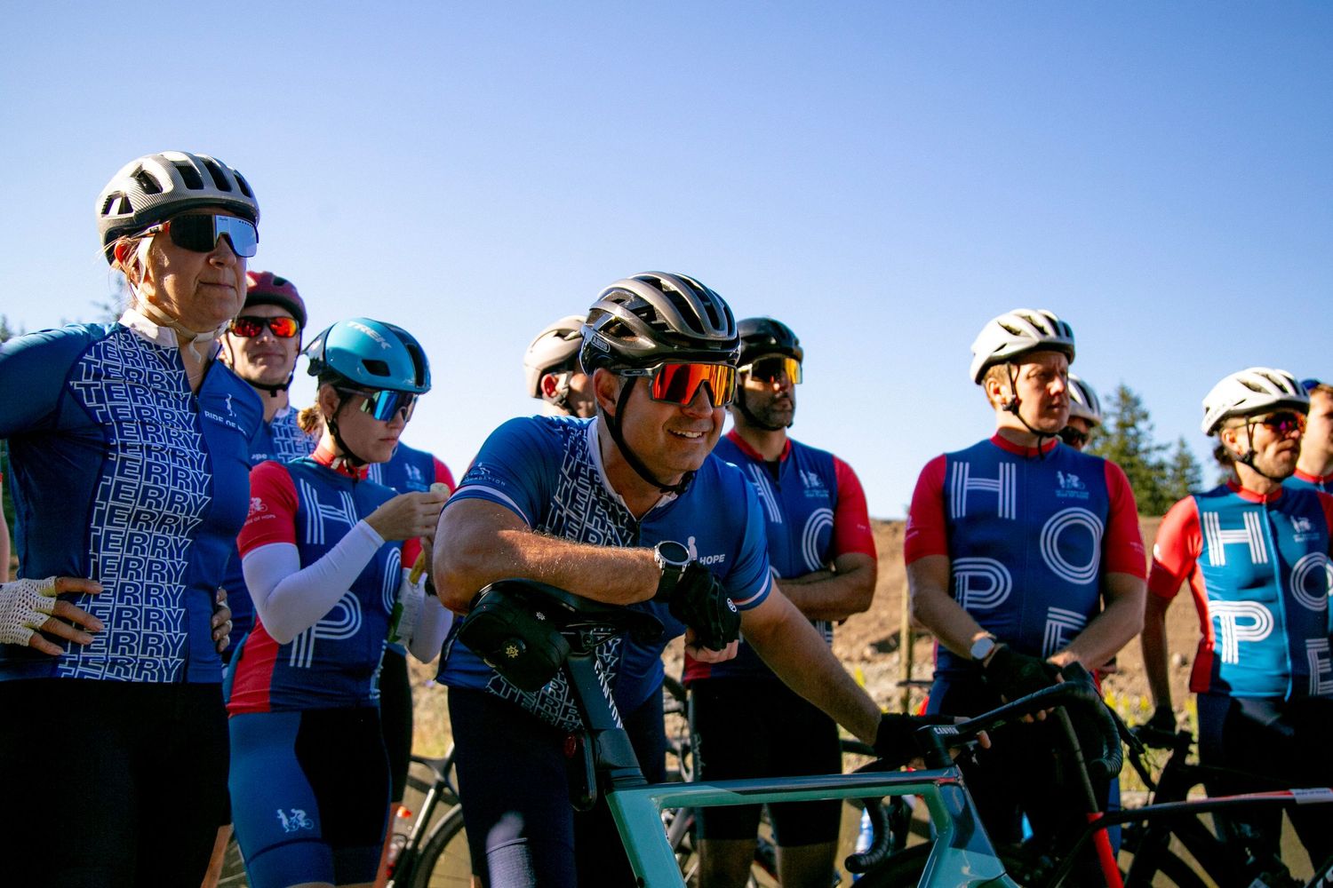 Group of cyclists in colorful jerseys gathered during daytime rest stop.