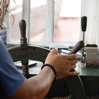 Hands operating vintage boat steering wheel controls in soft window light.