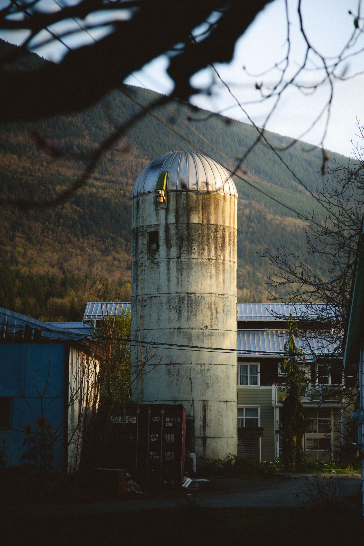 A weathered white grain silo stands next to farm buildings against a backdrop of forested mountains at dusk.