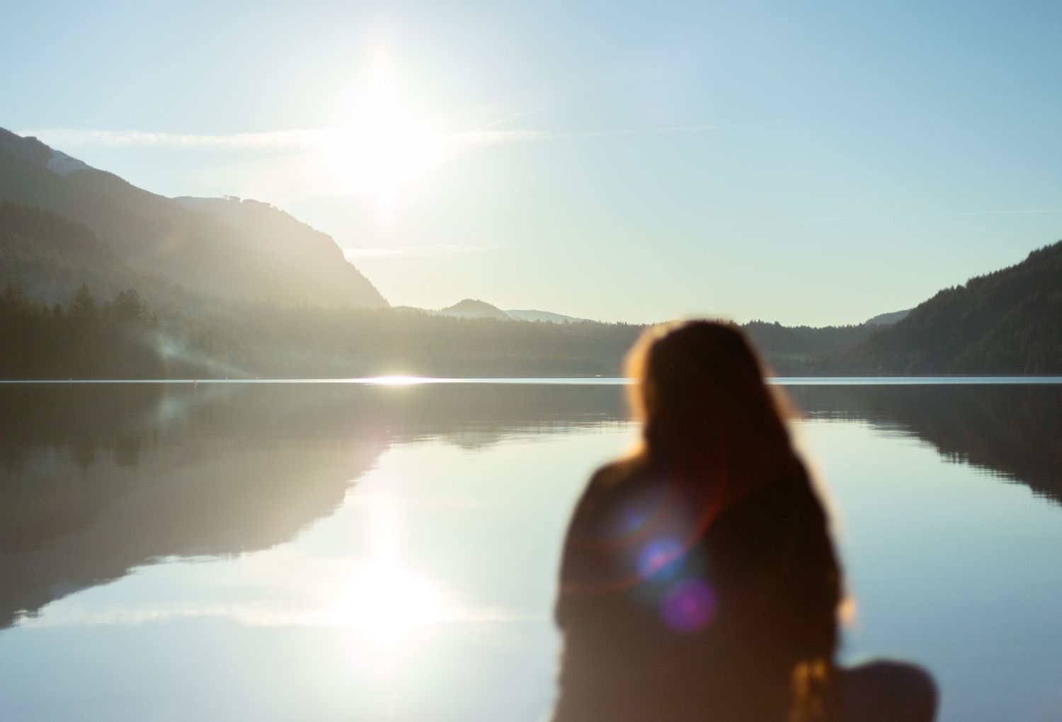 Silhouette figure watches sunrise over tranquil mountain lake with reflections on calm water.