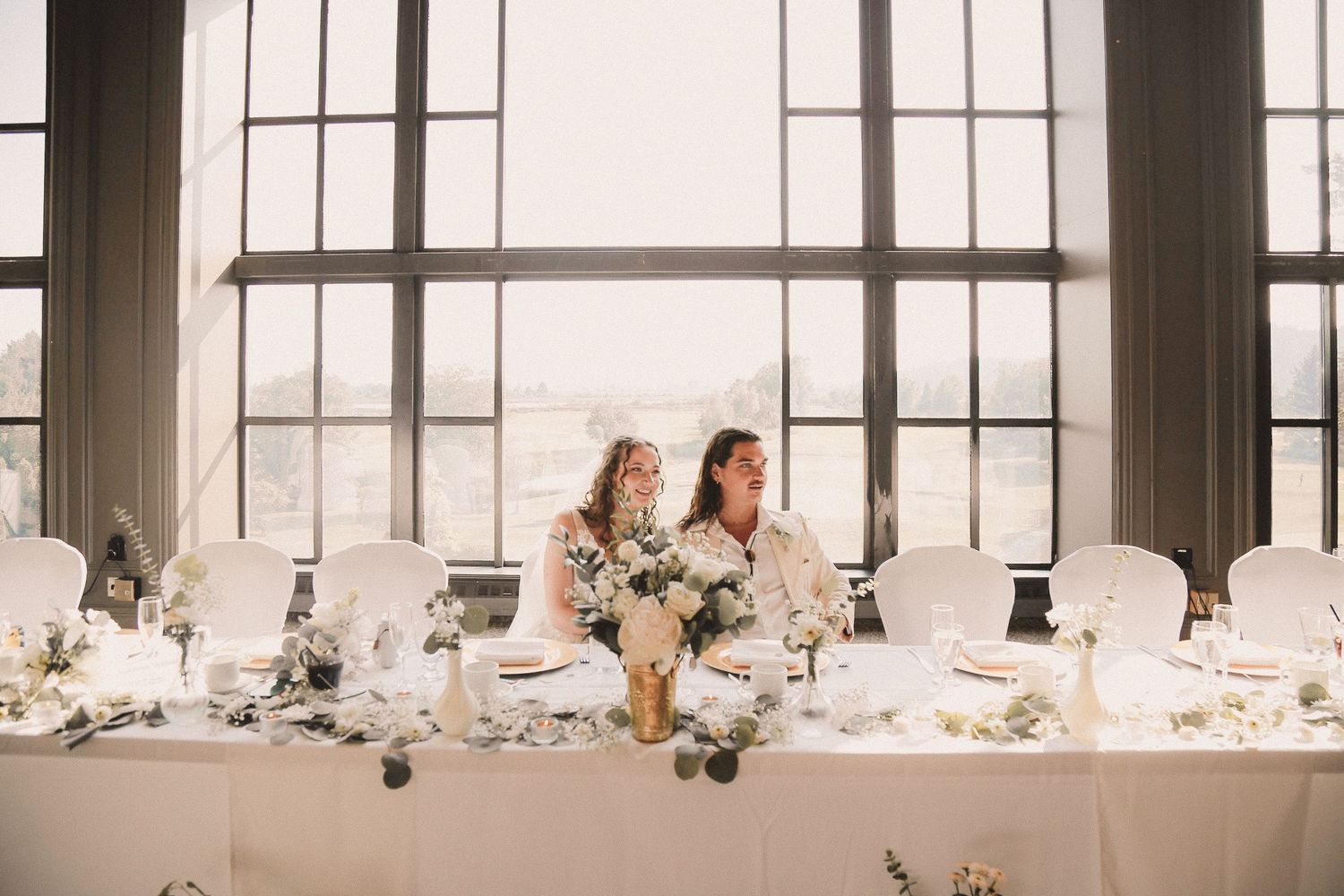 Elegant wedding reception table setting by large windows with white linens and floral arrangements.