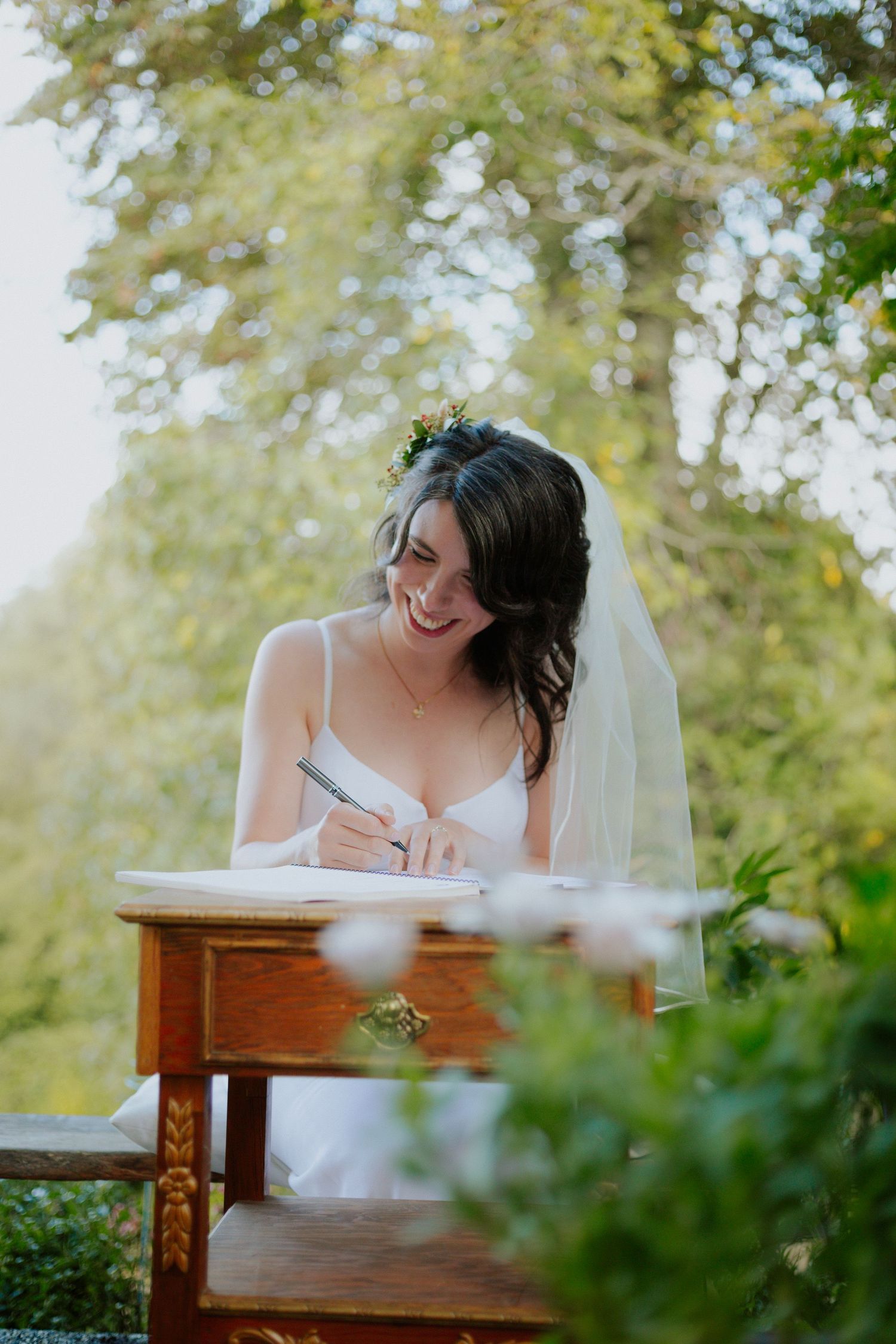 A bride in white dress signs documents at wooden desk in outdoor garden setting.