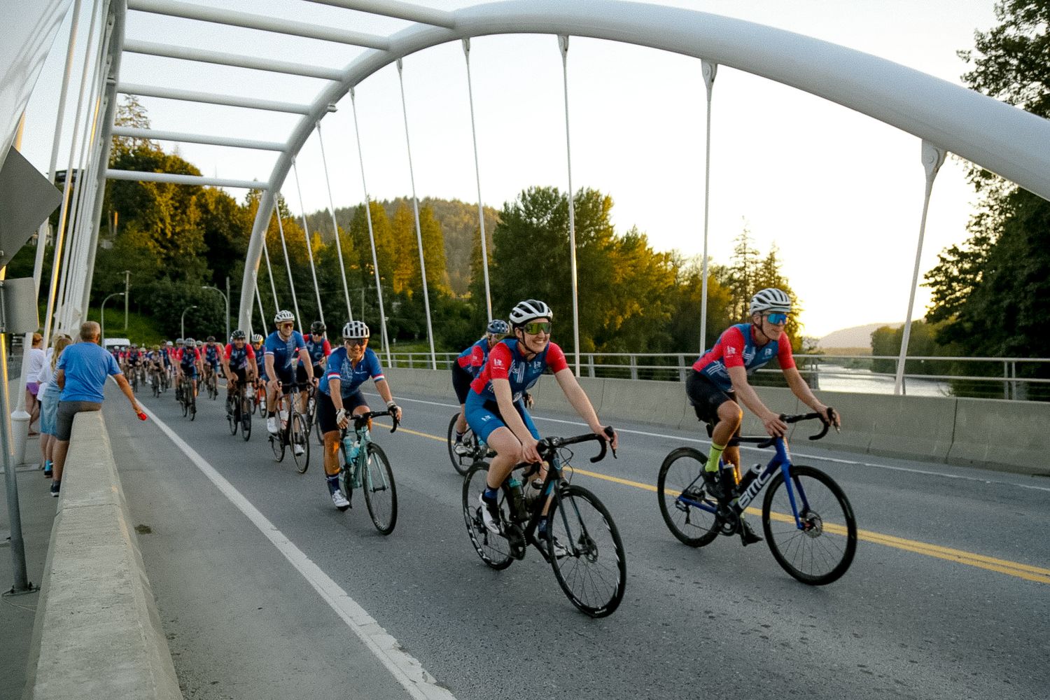 Peloton of cyclists crossing a white arched bridge during sunset.