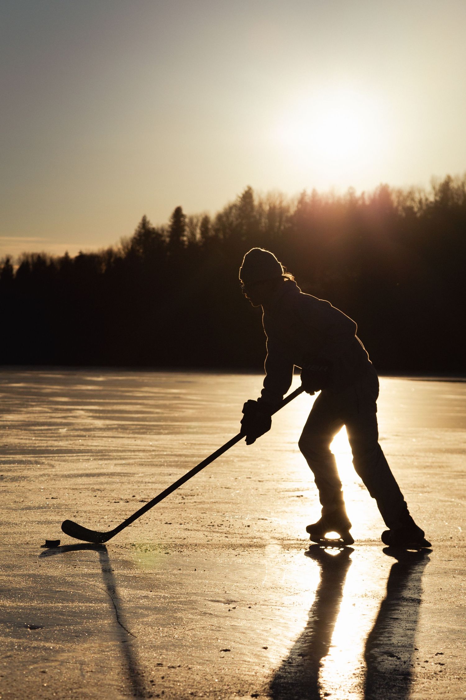 Silhouette of ice hockey player practicing on frozen lake at sunset with forest backdrop and glowing sunlight.