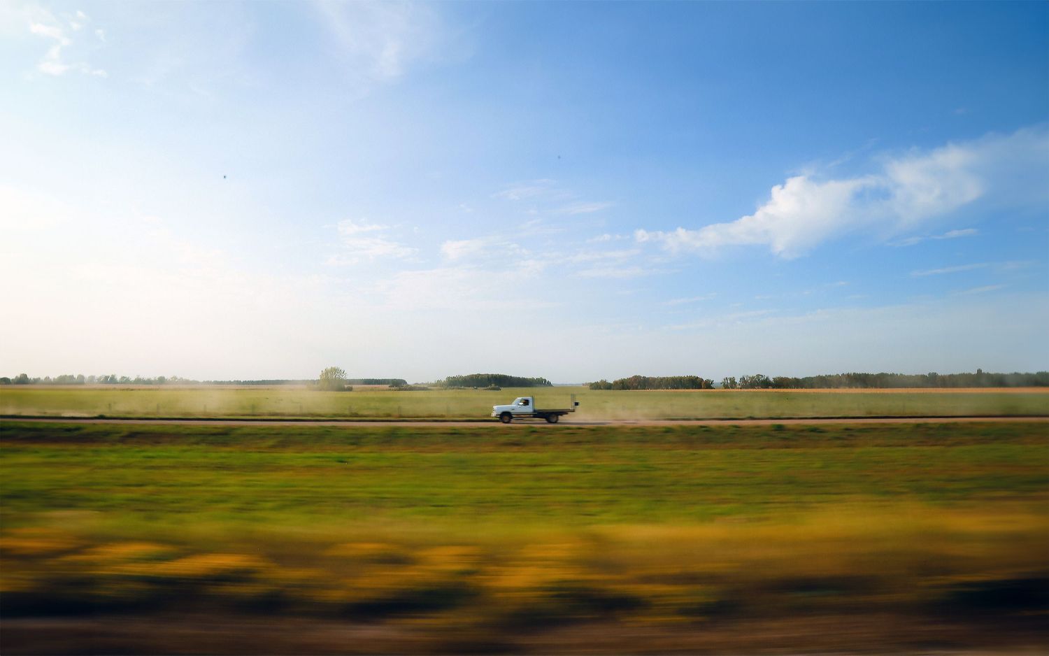 A car drives through an open grassland under a vast blue sky.