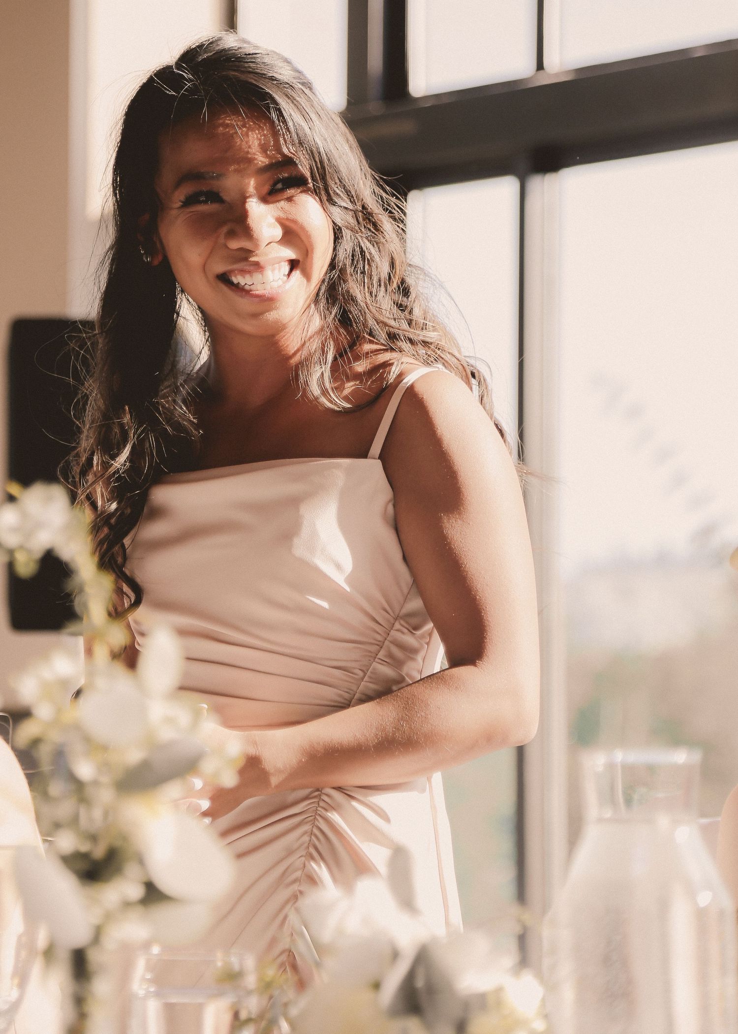 Woman in blush dress poses by window with floral arrangements.