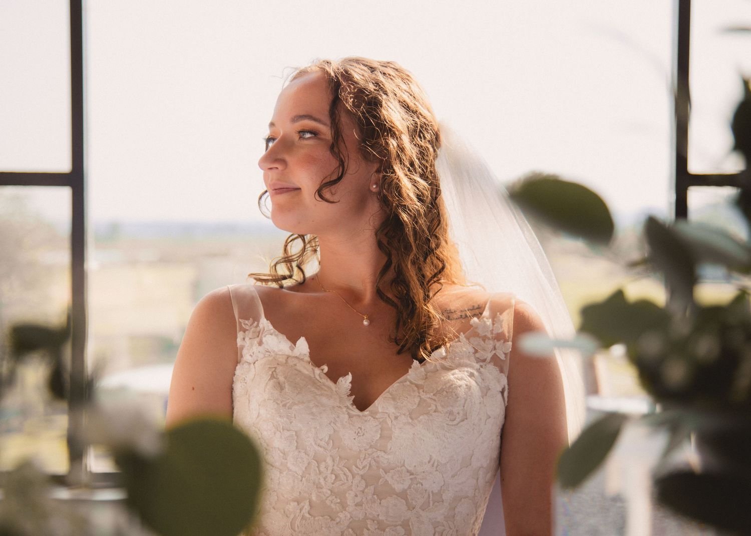 Bride in lace wedding dress gazes to the side in sunlit window setting.