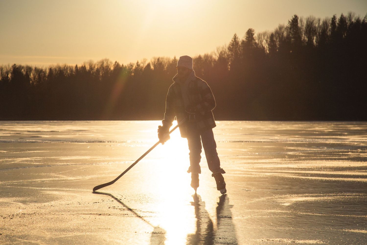 Silhouette of hockey player skating on frozen lake at sunset with stick in hand against golden sky.