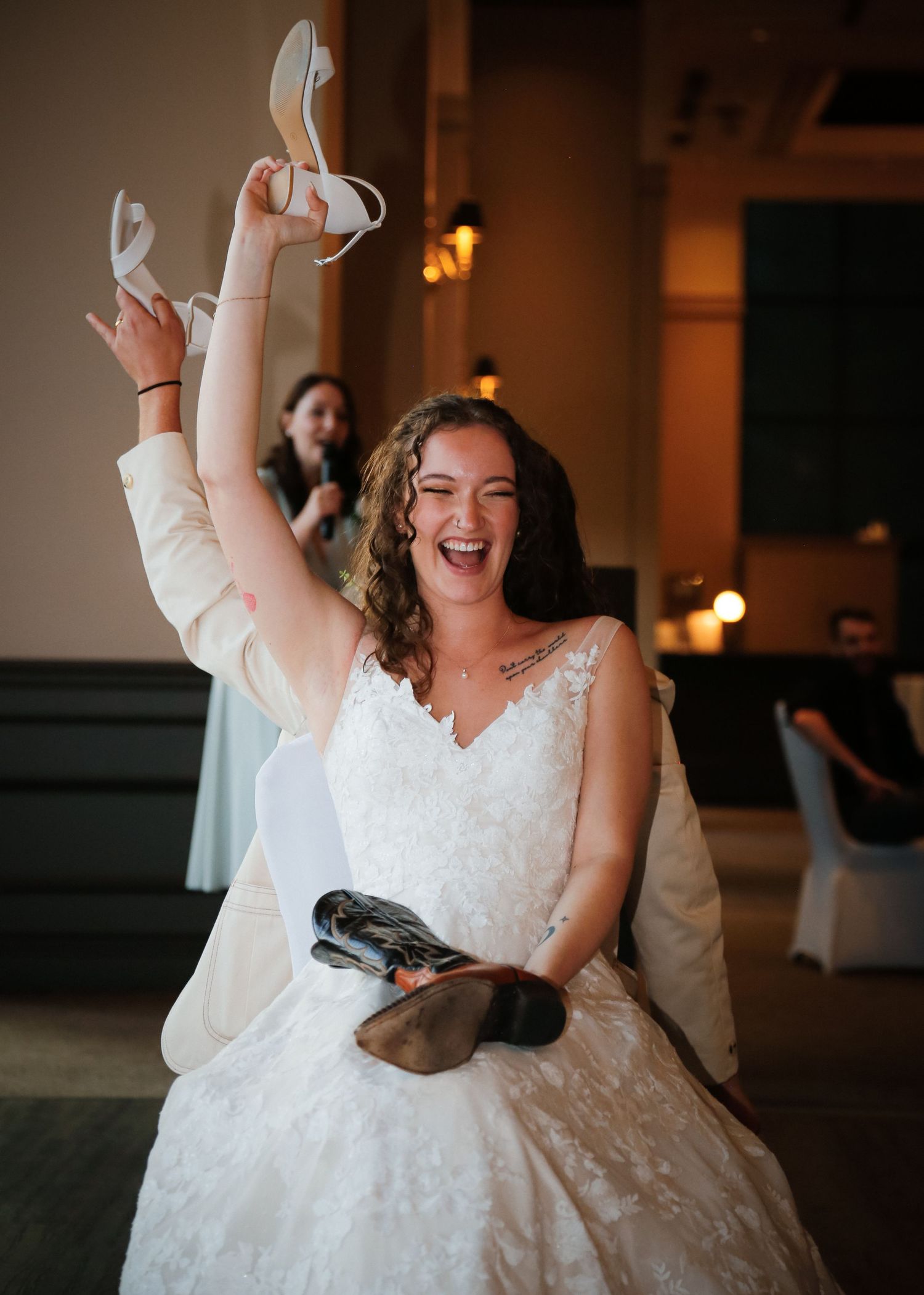 A joyful bride in a white lace dress holds up a shoe while sitting at her wedding reception.