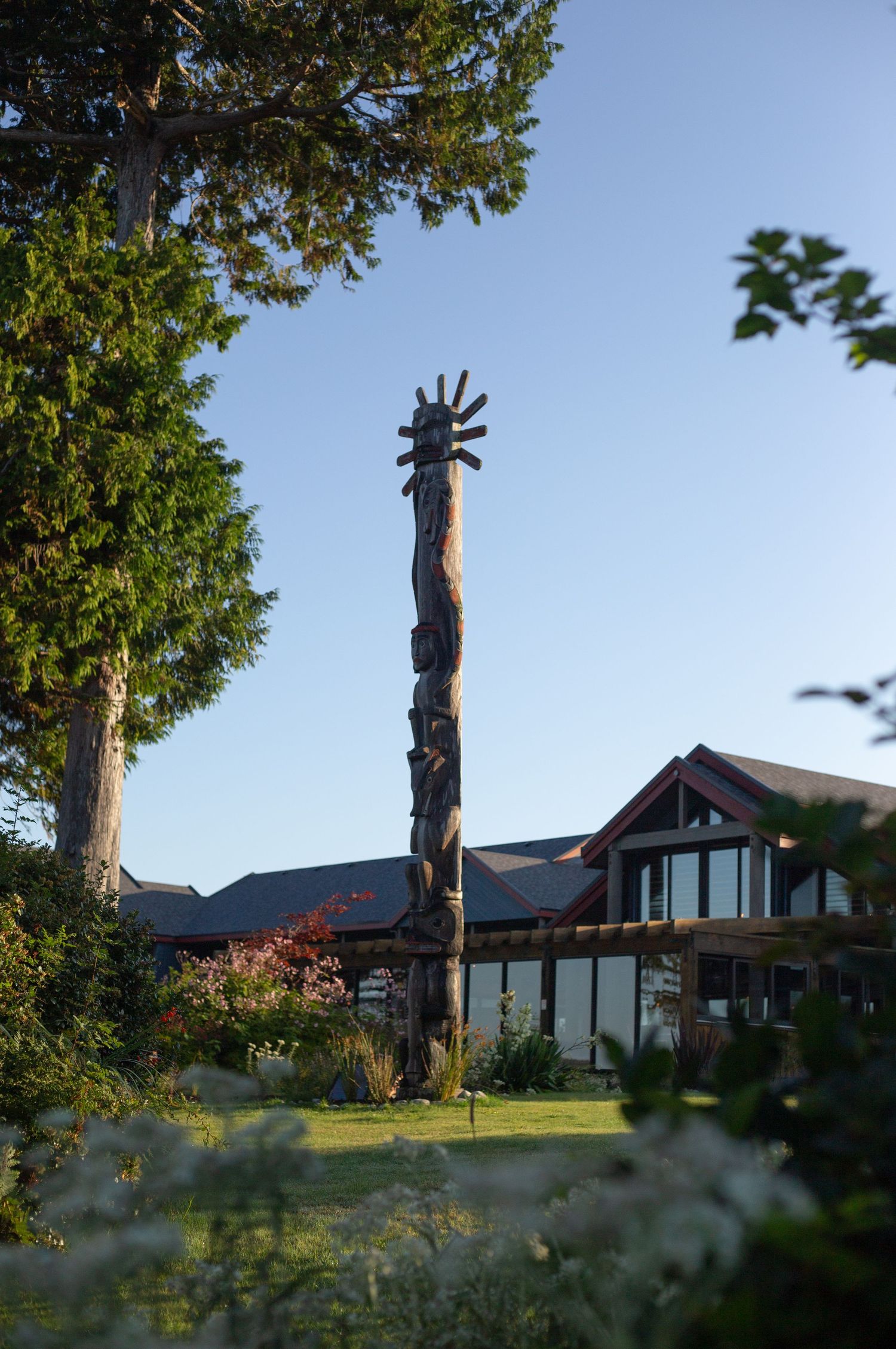 Tall carved wooden totem pole stands in front of house with trees and blue sky.