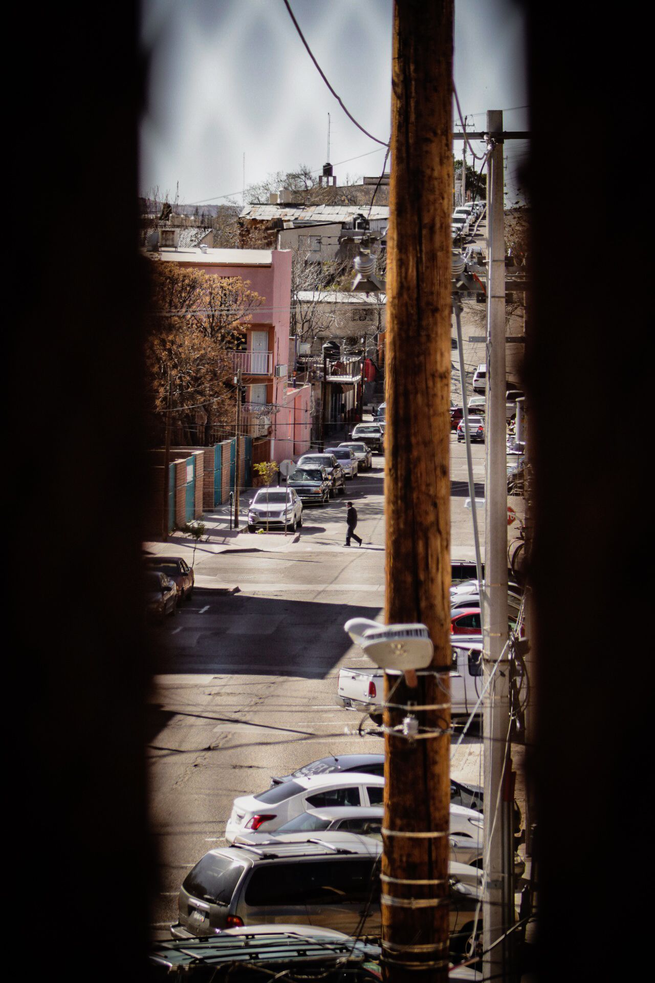 A narrow urban street viewed through wooden poles with parked cars and buildings on both sides.