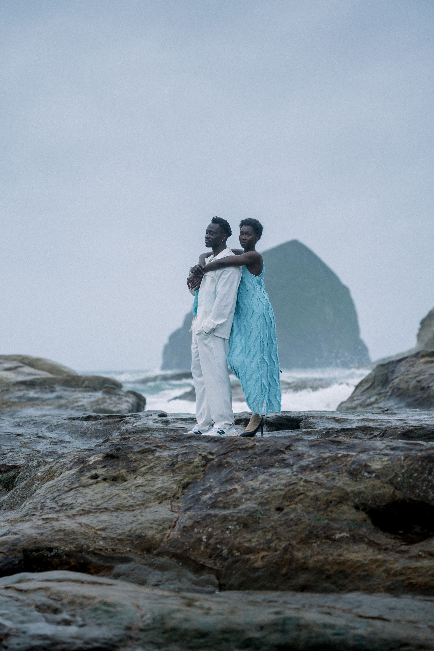 Two people in flowing garments stand together on rocky cliffs with a large sea stack visible in the background.