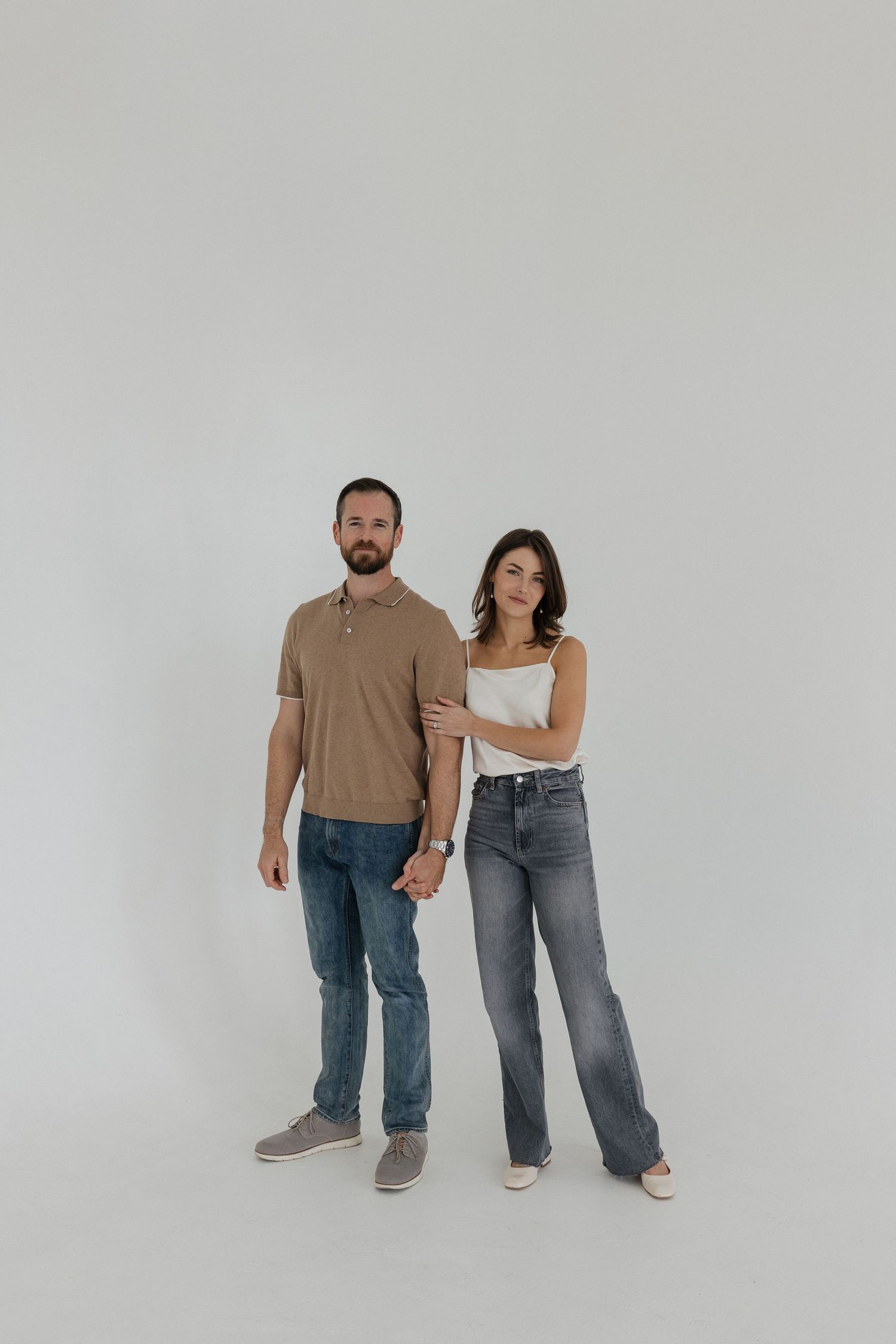 Couple in casual attire standing against white backdrop in minimalist studio setting.