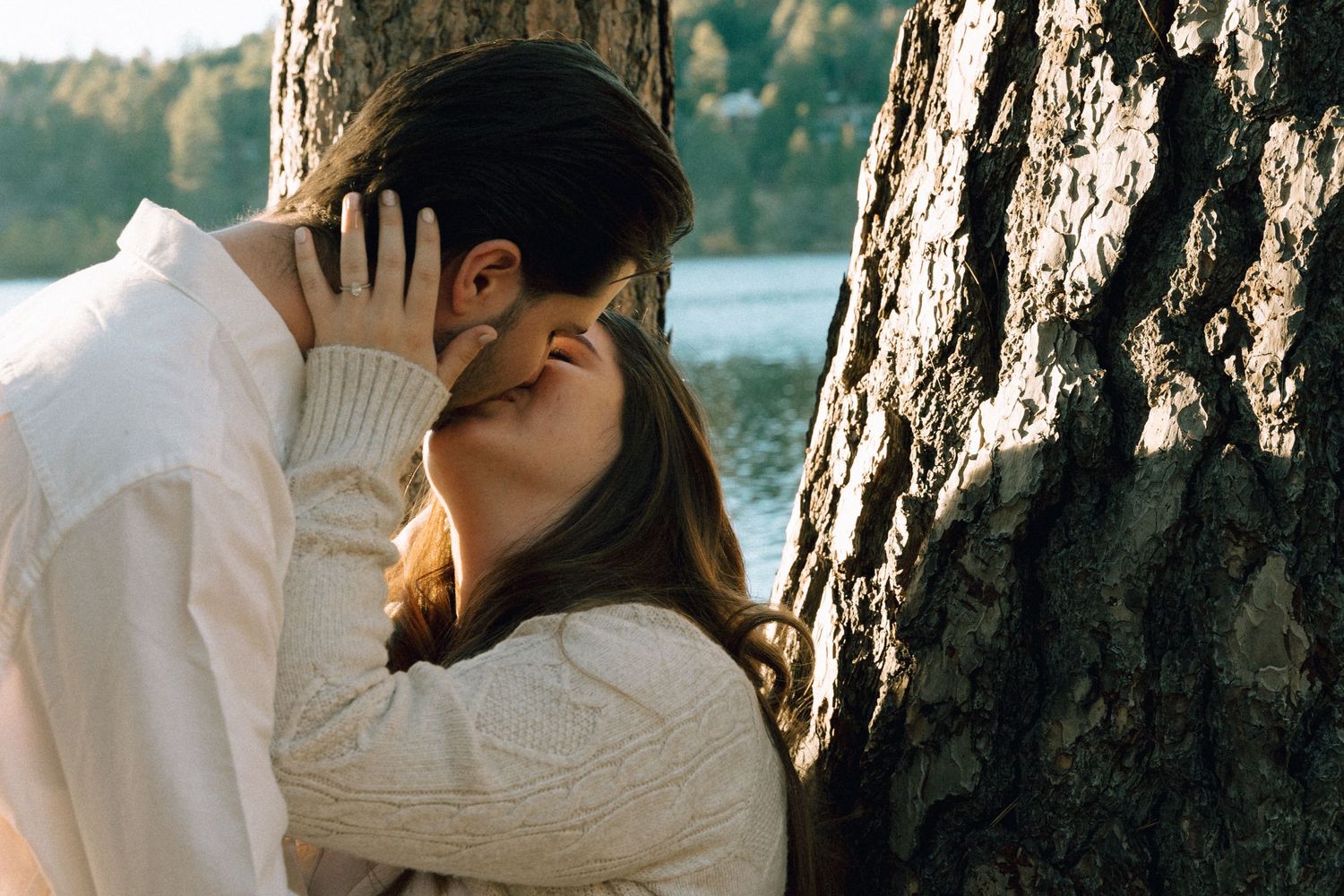 Romantic moment between couple leaning against tree trunk by lakeside.