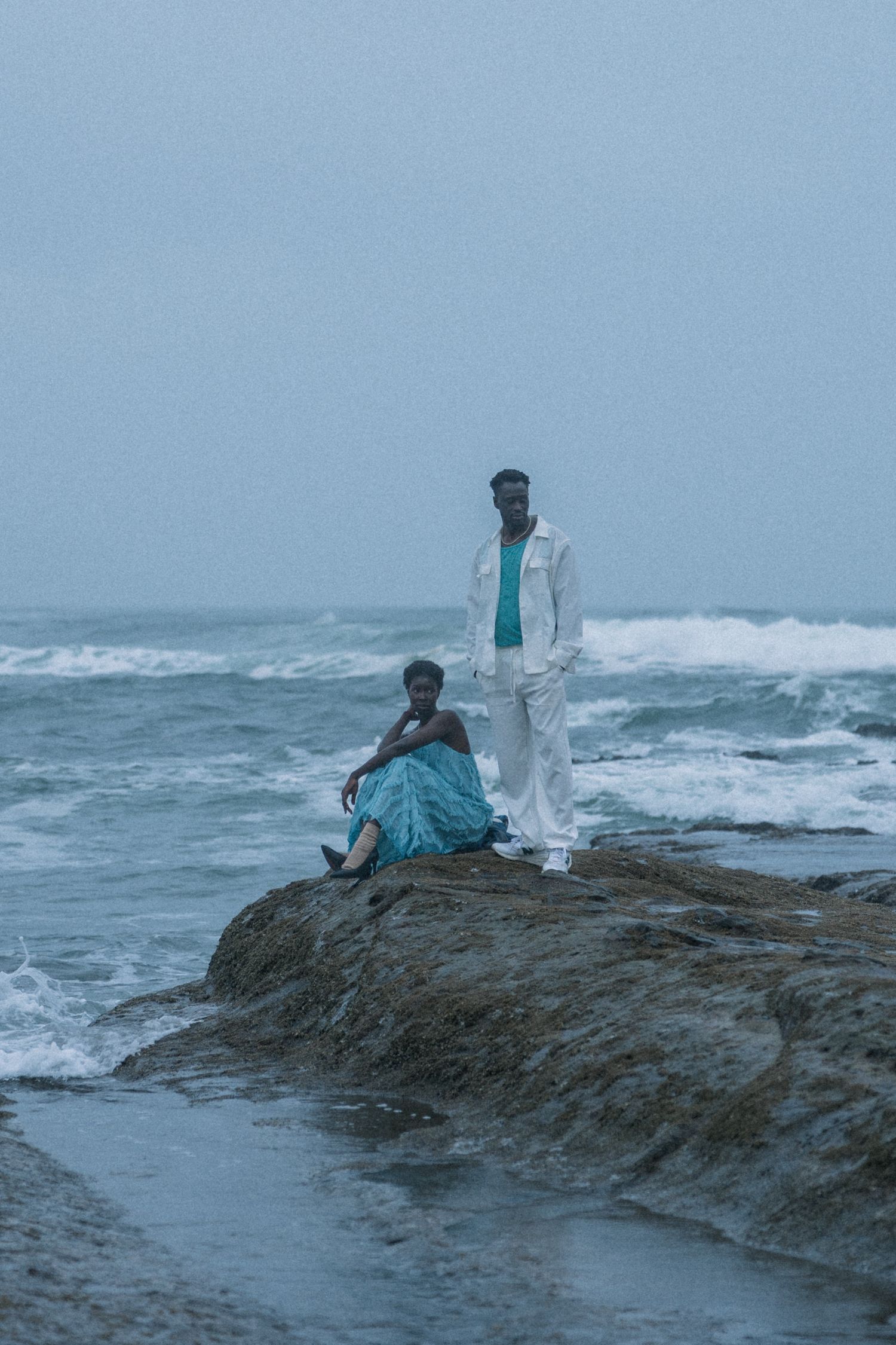 Two people in contrasting outfits rest on oceanside rocks as waves crash behind them.