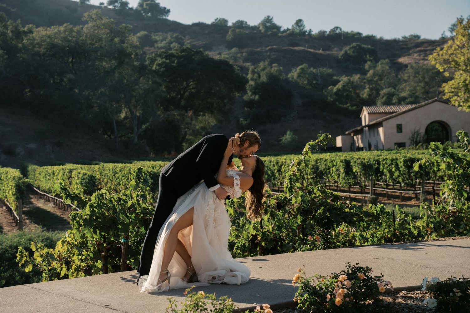 Newlyweds share an intimate moment on a paved path overlooking scenic vineyard rows.