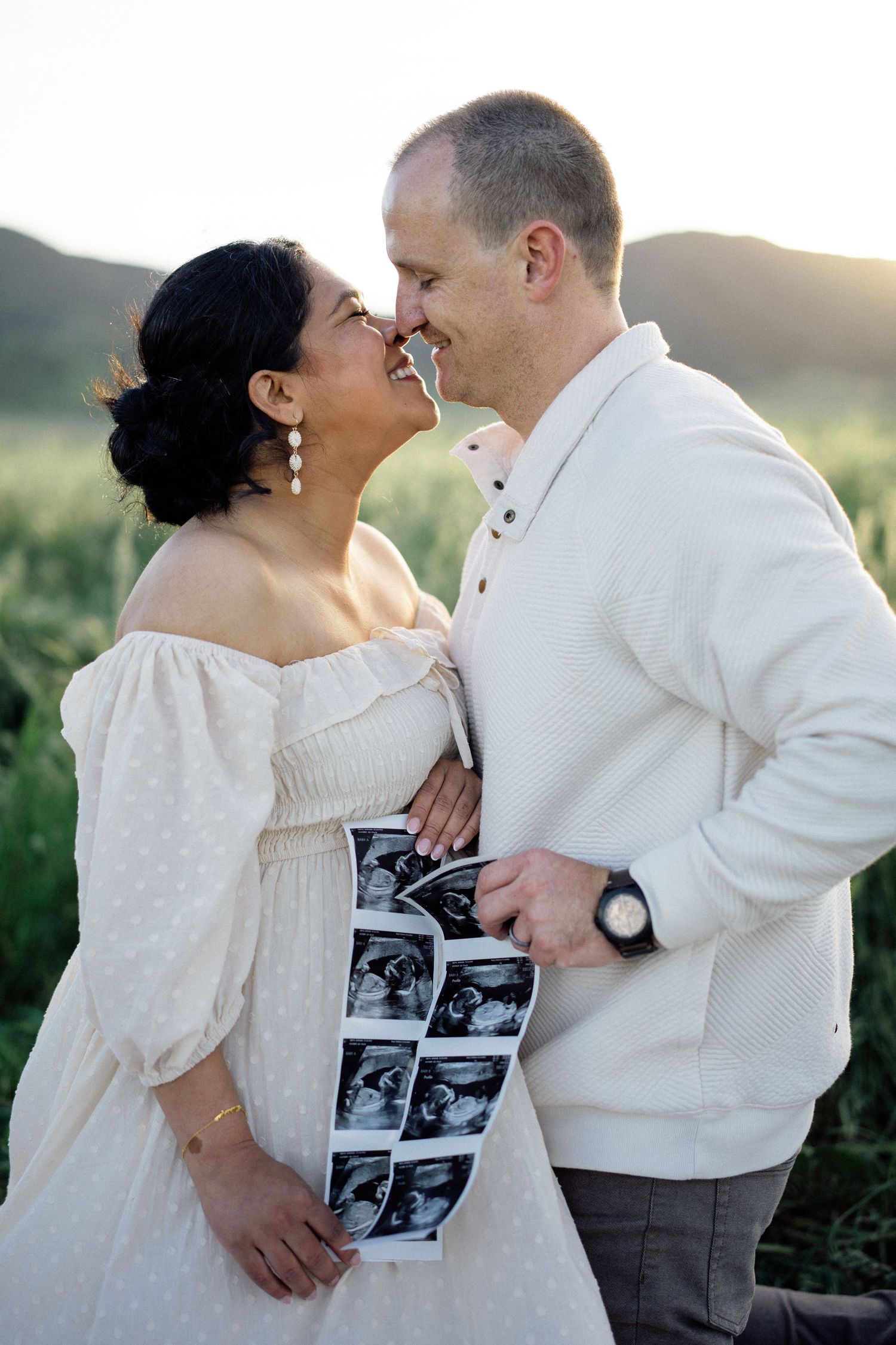 A couple in white outfits share an intimate moment while holding ultrasound photos outdoors at sunset.