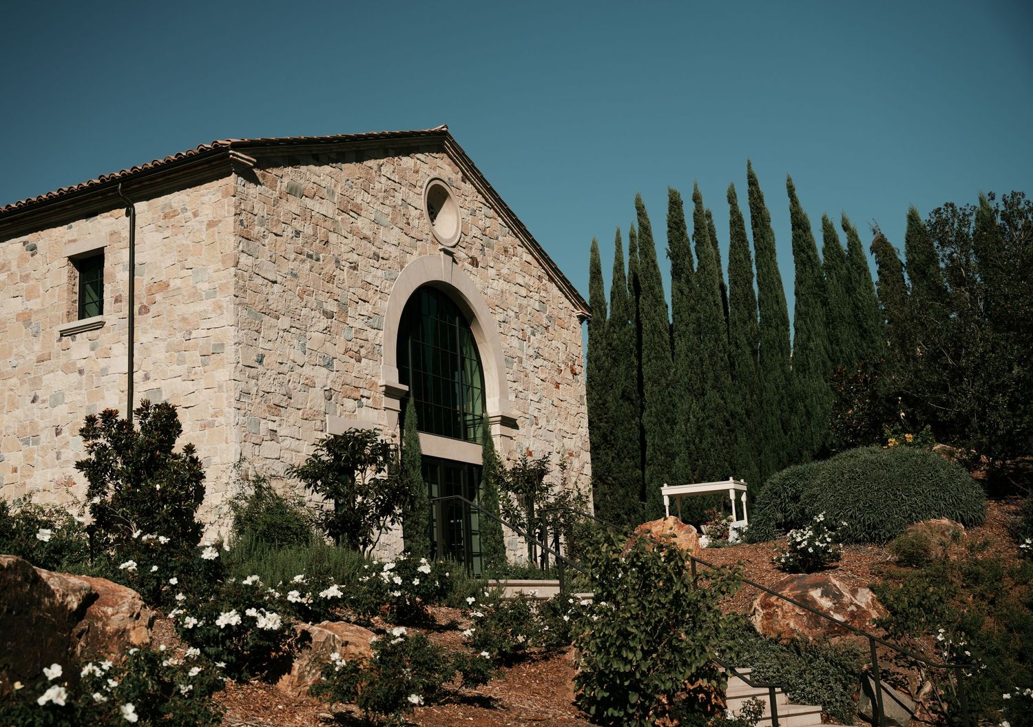 Rustic stone villa with arched entrance surrounded by cypress trees and Mediterranean landscaping.