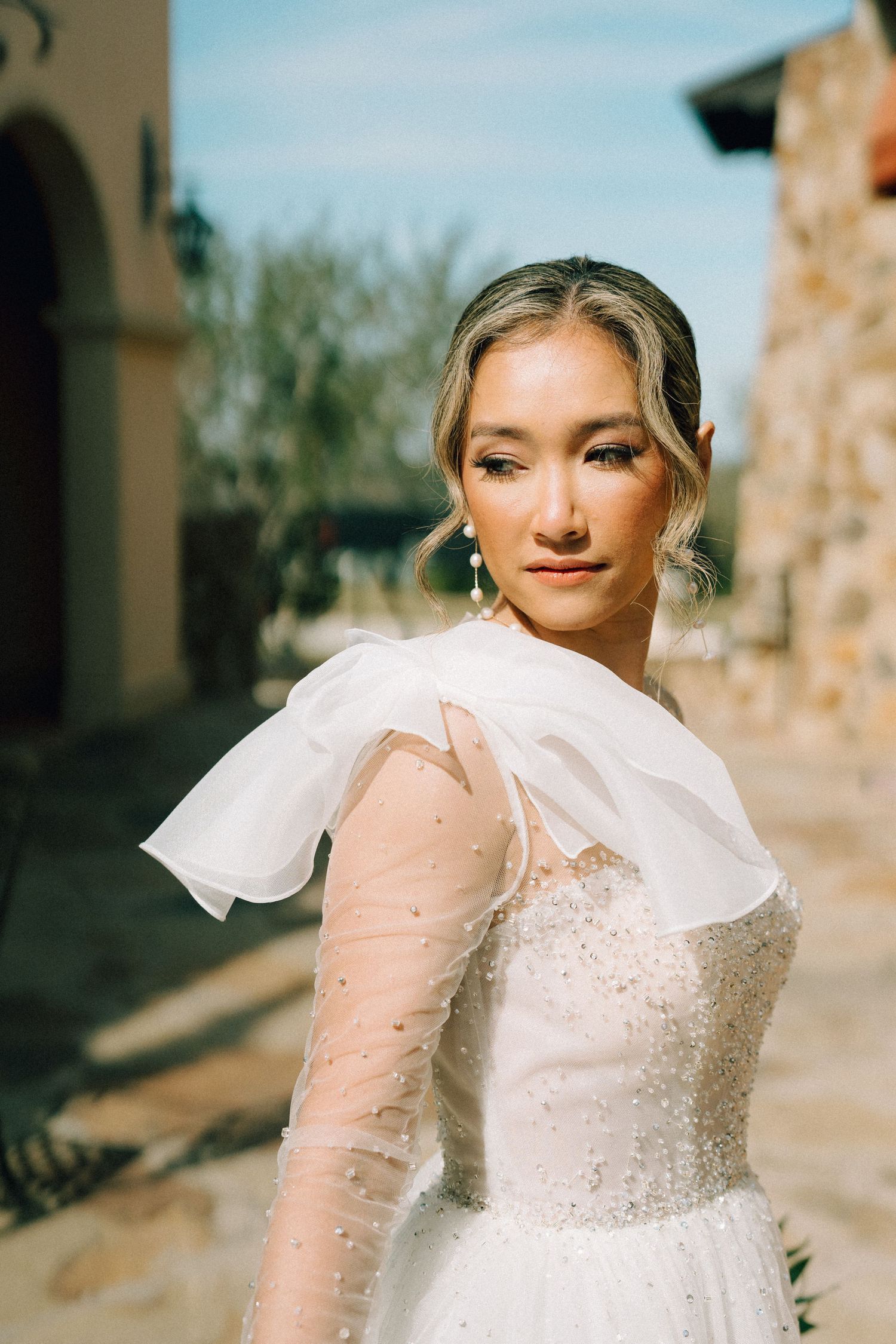 A close-up portrait of a bride wearing a white dress with sheer sleeves against a stone backdrop.