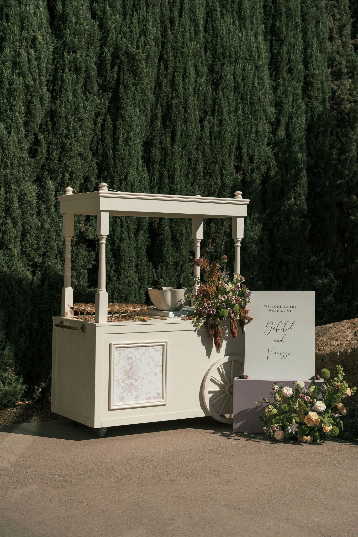 White wedding dessert cart setup against tall cypress trees with floral decorations.