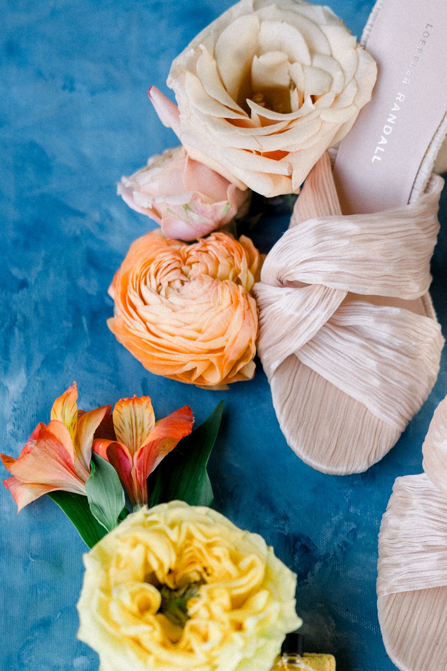 Wedding shoes and flowers arranged on a blue background featuring roses and ranunculus blooms.